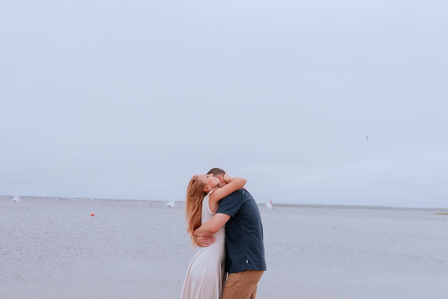 Couple sharing a romantic kiss on a sandy beach with calm ocean waters and overcast sky in the background.