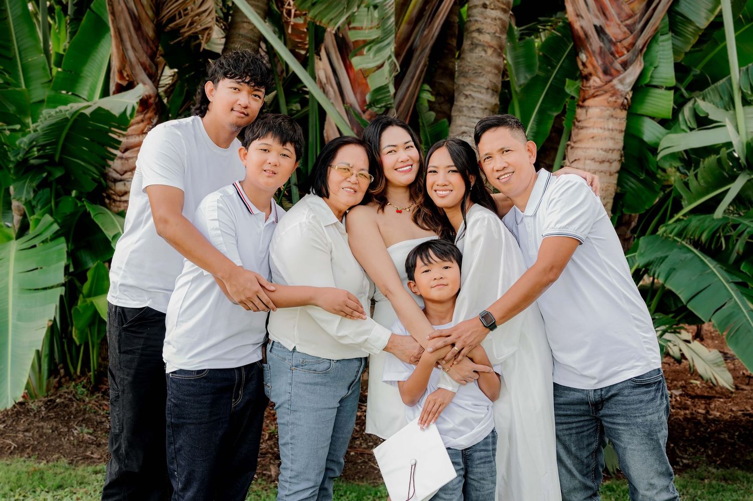 Happy family of seven posing together outdoors in white outfits surrounded by tropical palm trees and greenery.