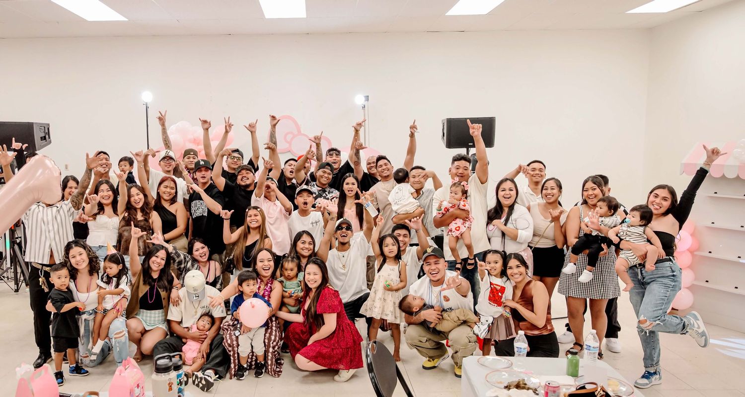 Large group of smiling people celebrating at a party with pink balloons and decorations in a bright white room.