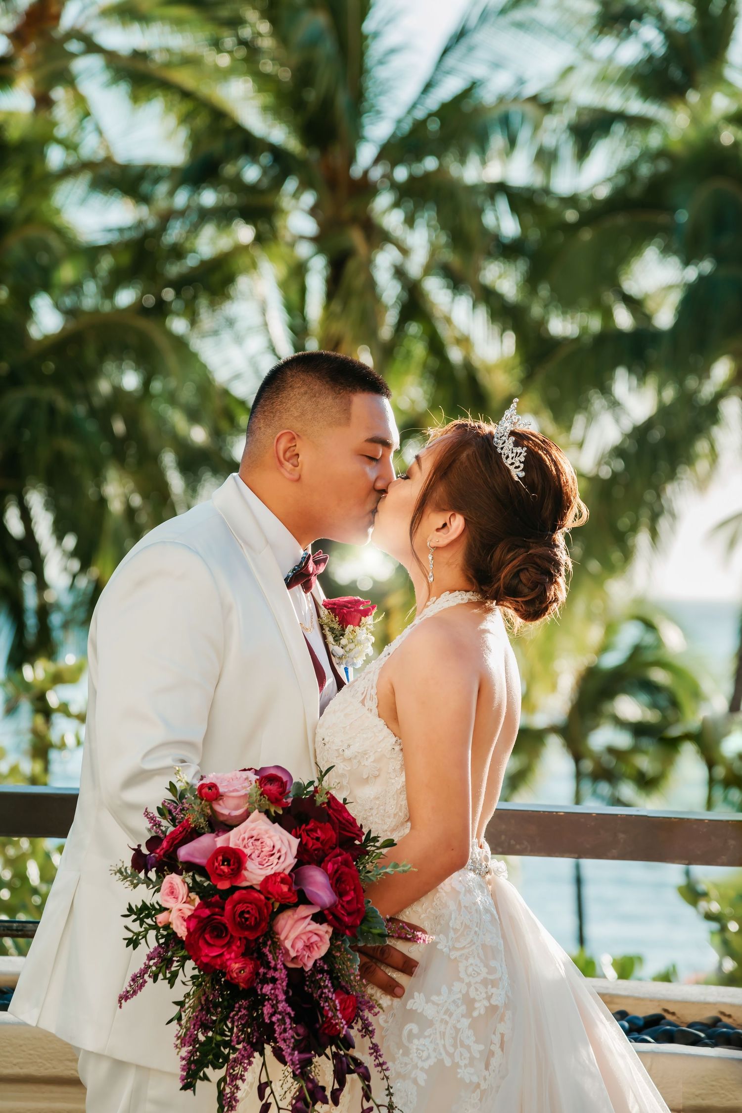 A romantic wedding couple shares a kiss on a tropical balcony with palm trees and a stunning pink and red bouquet.