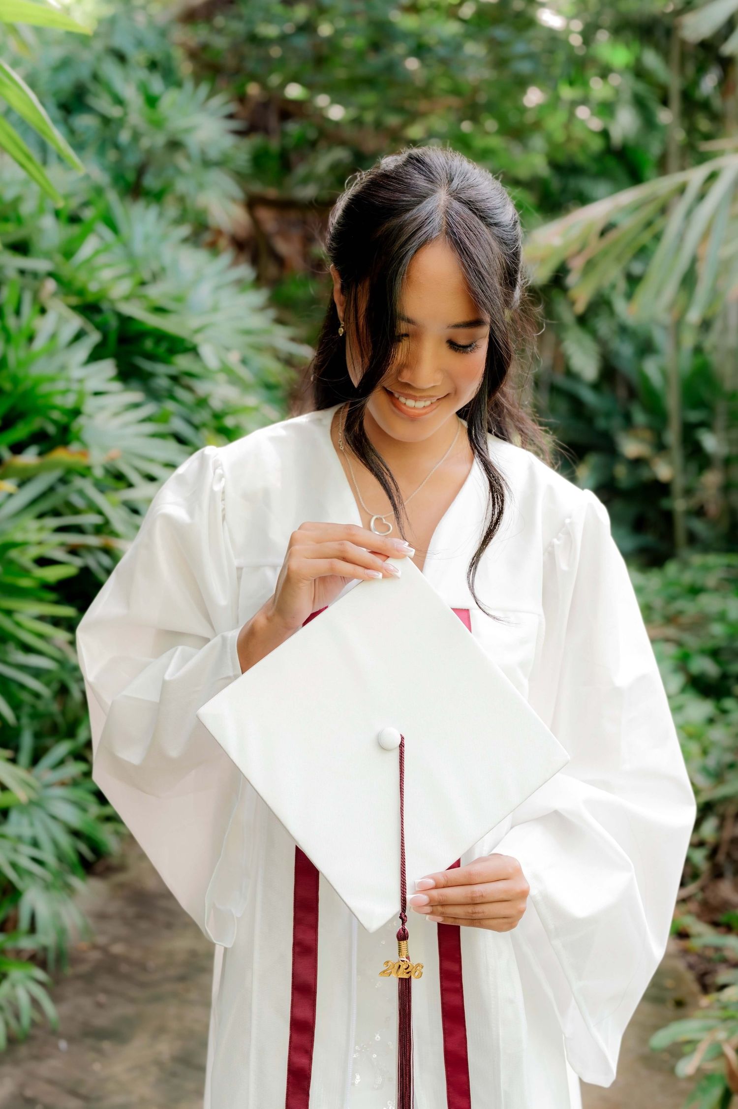 Smiling young woman in white graduation cap and gown holding her mortarboard outdoors among lush green tropical foliage.