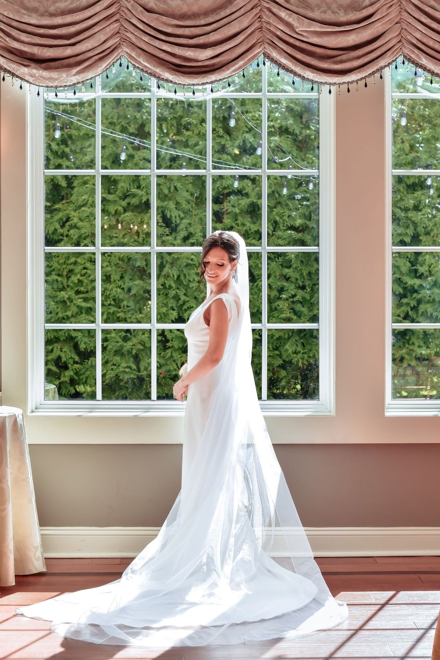 A bride in an elegant white wedding gown with cathedral veil poses in front of tall windows with draped valances.