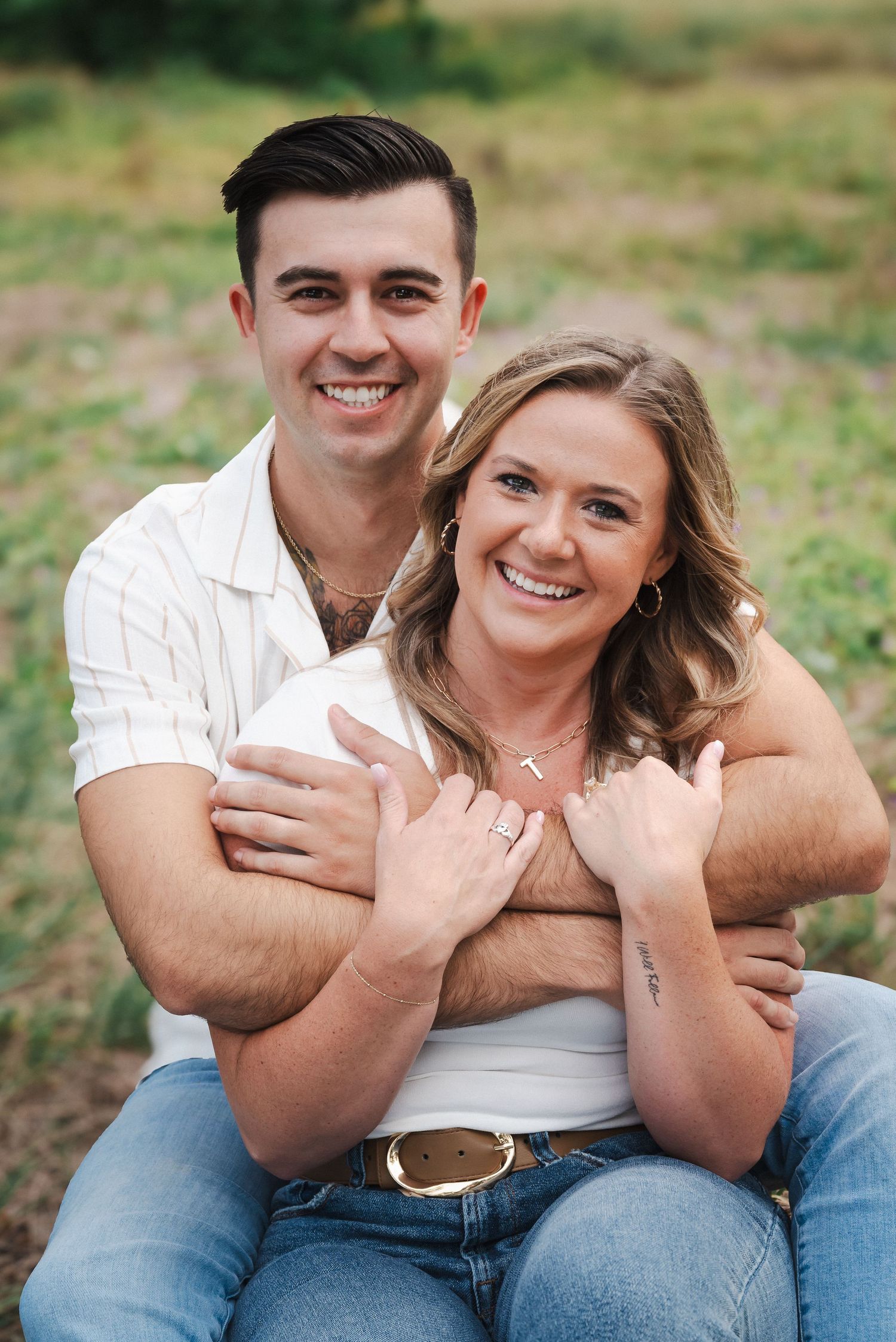 Young couple embraces while sitting outdoors in casual clothing on a sunny day in a grassy field.
