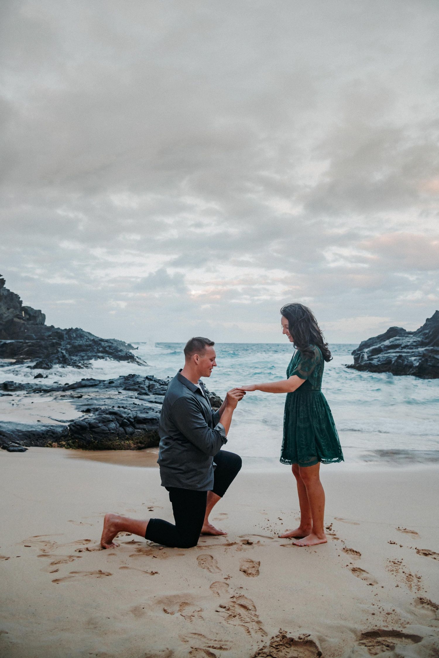 Man proposes to woman on one knee on a sandy beach with rocky shoreline and dramatic cloudy sky backdrop.