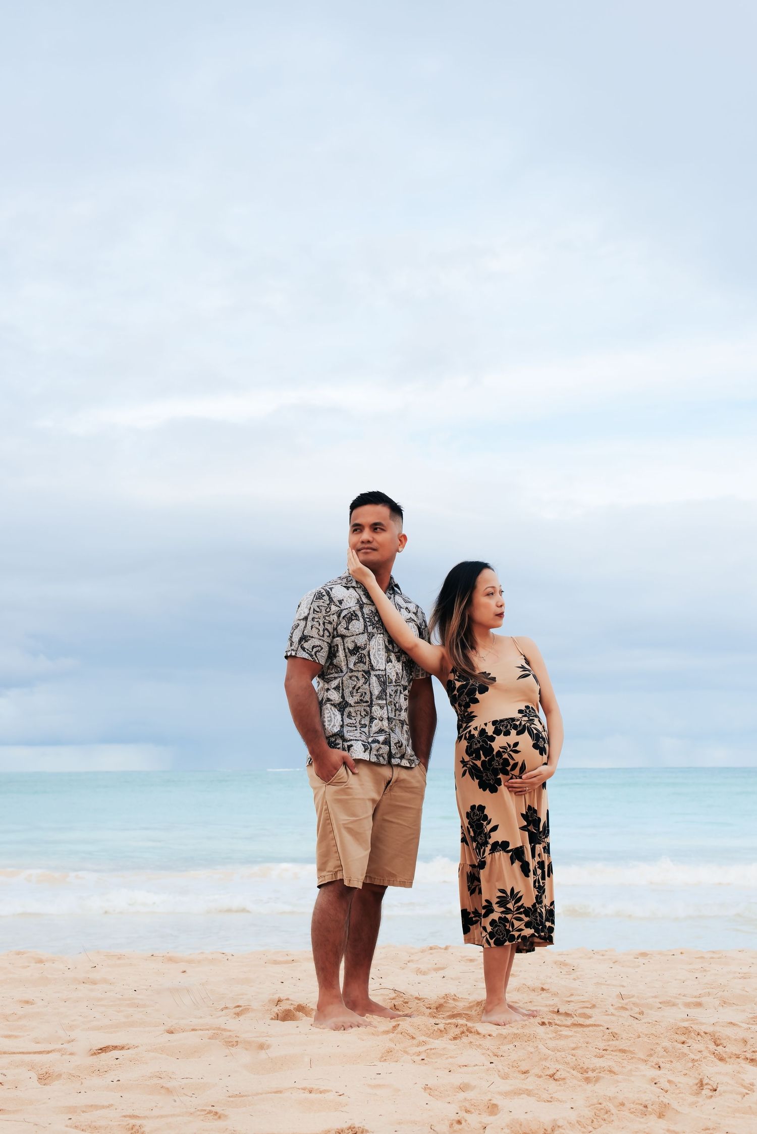 Pregnant couple standing on a sandy beach, man in floral shirt and shorts, woman in black floral dress cradling baby bump.