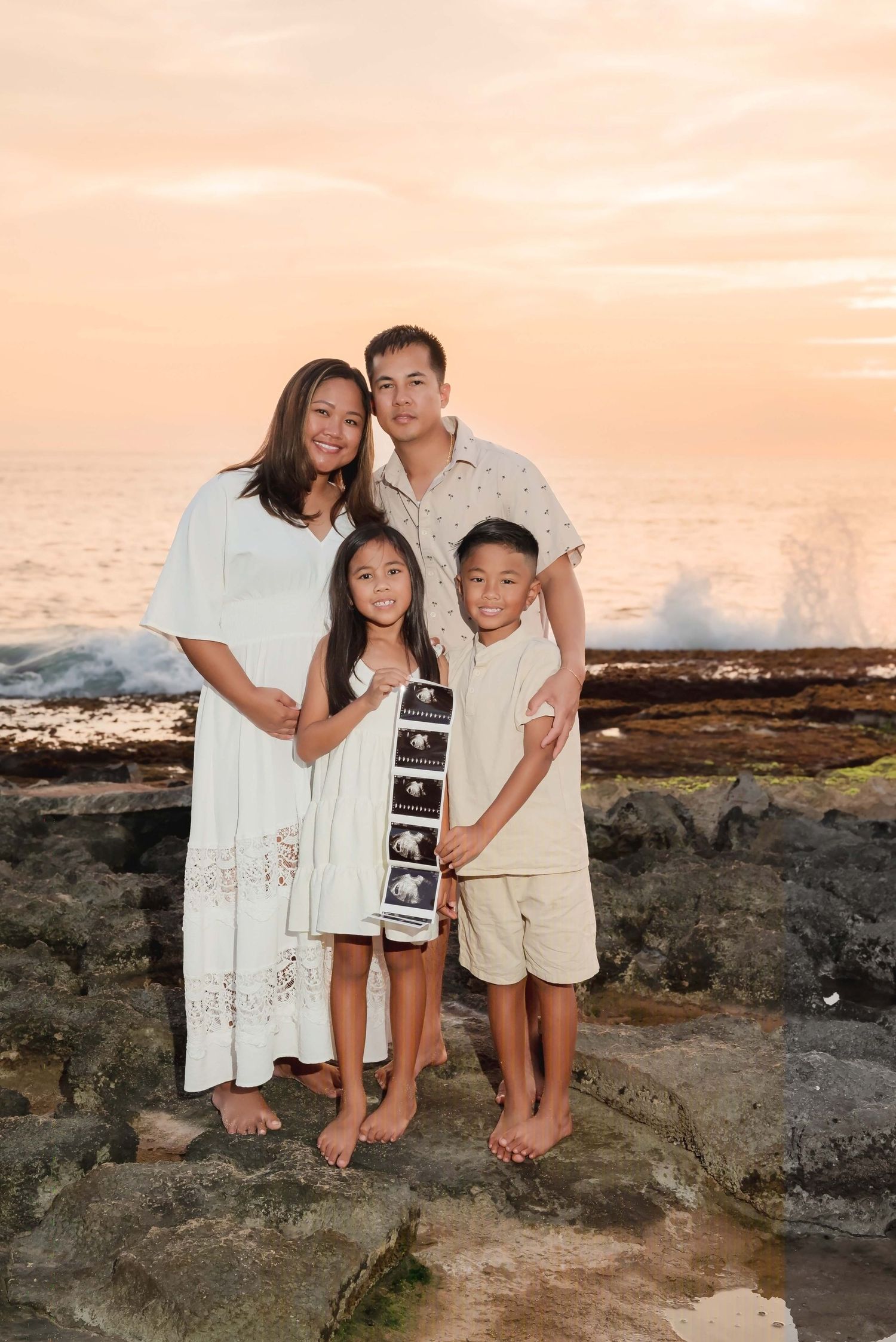 Family of four poses on rocky shore at sunset, mother visibly pregnant, children holding ultrasound photos by ocean waves.