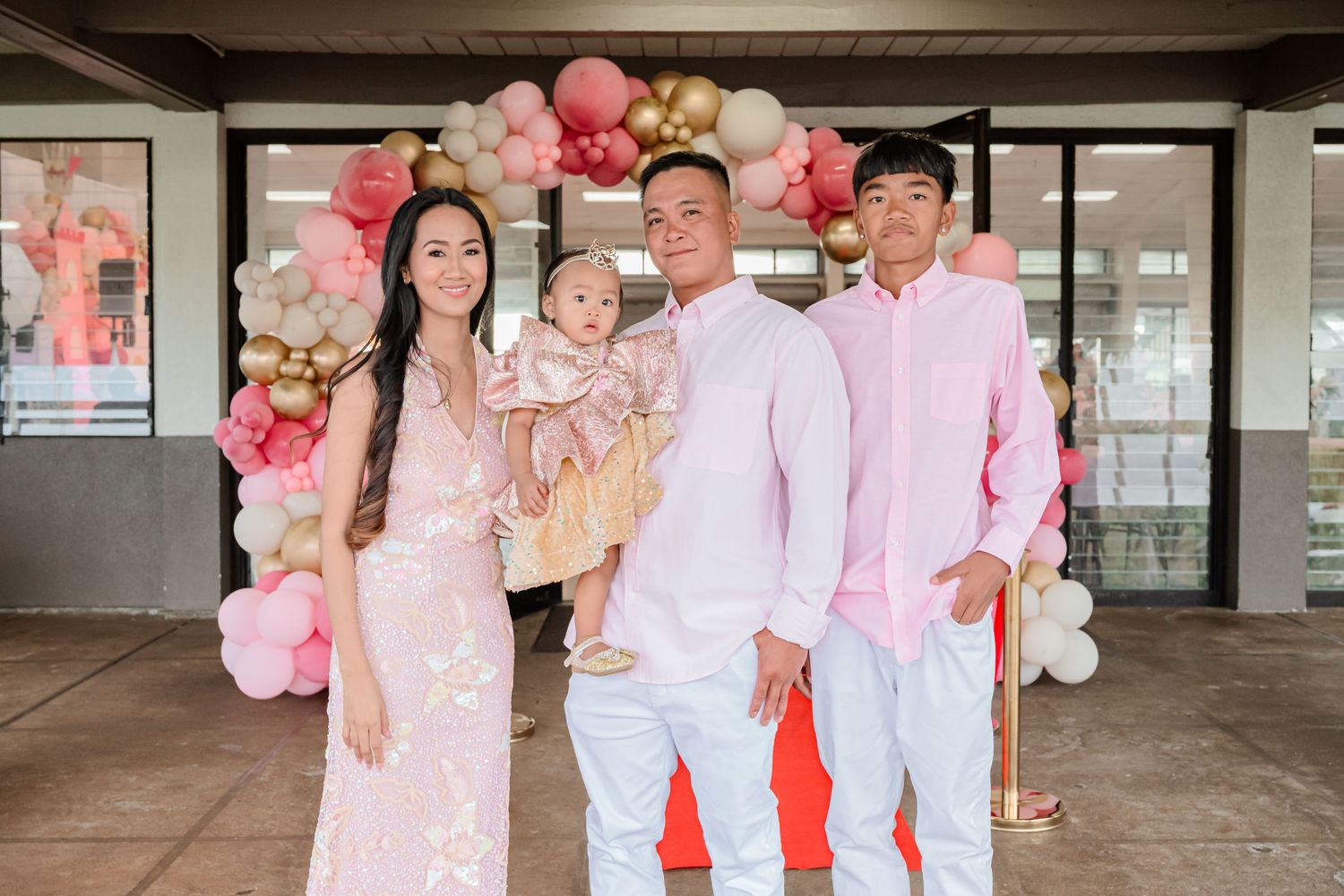 Family of four posing in pink outfits at a festive balloon backdrop celebration event.