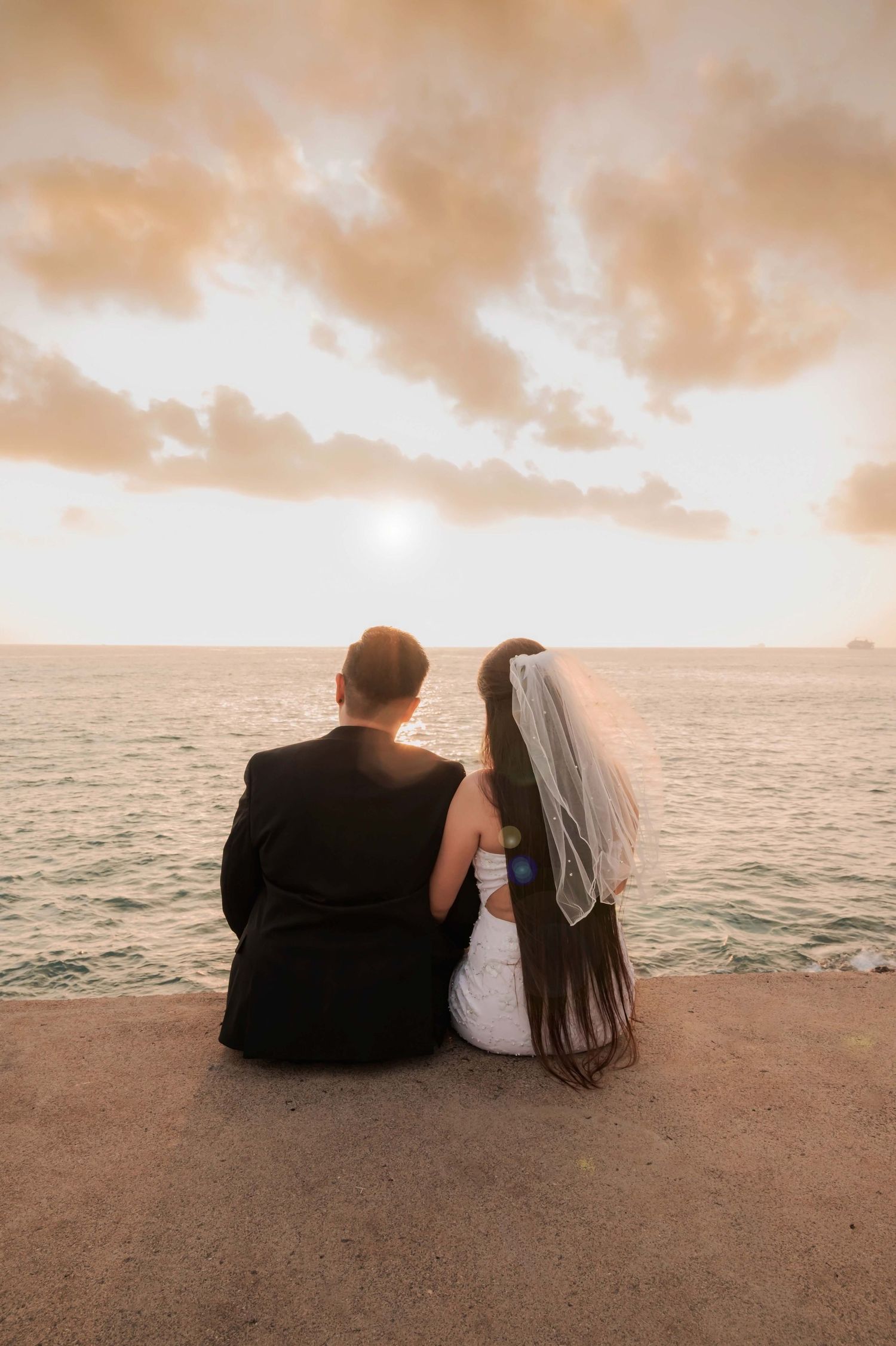 Newlywed couple in wedding attire sitting on a seaside wall watching a romantic golden sunset over the ocean.