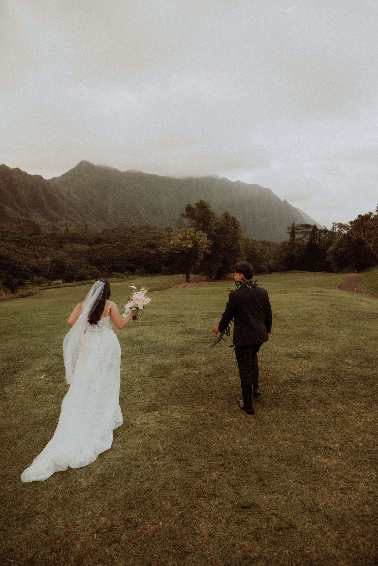 A couple in wedding attire walk across a grassy field with dramatic mountain peaks in Hawaii under moody skies.