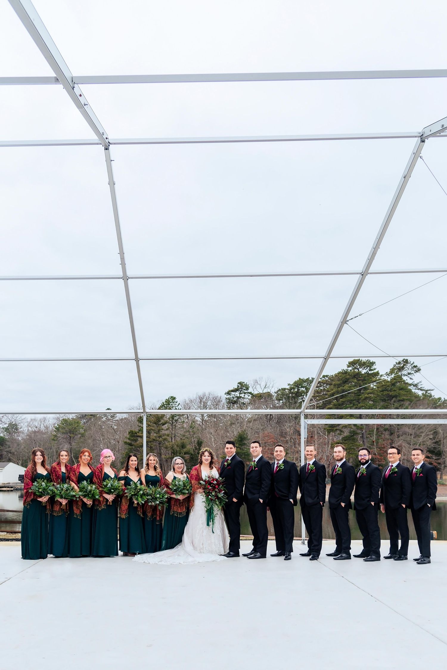 A wedding party in emerald green and black formal attire poses under a white tent structure.