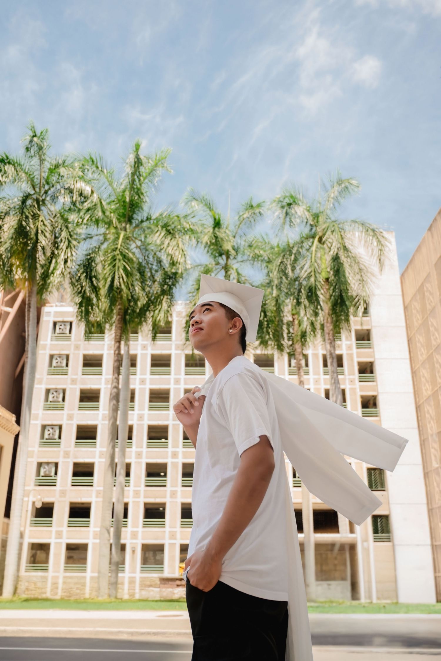 Young man in white graduation cap and gown poses outdoors near palm trees and a multi-story building on a sunny day.