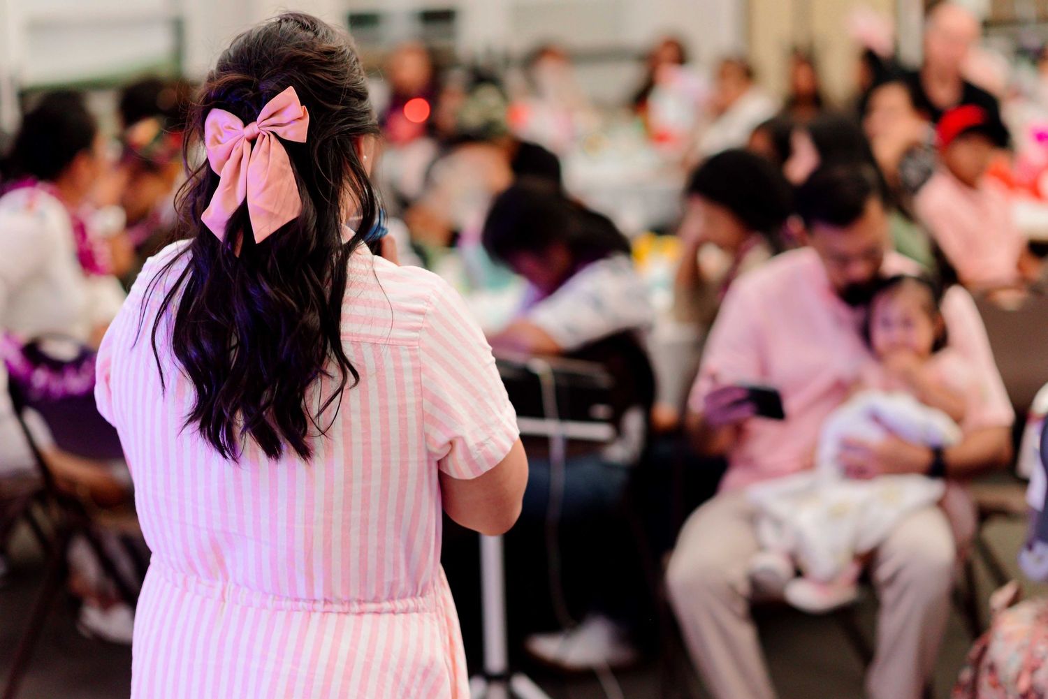 Woman with pink bow in dark hair wearing pink dress at a crowded indoor gathering event.