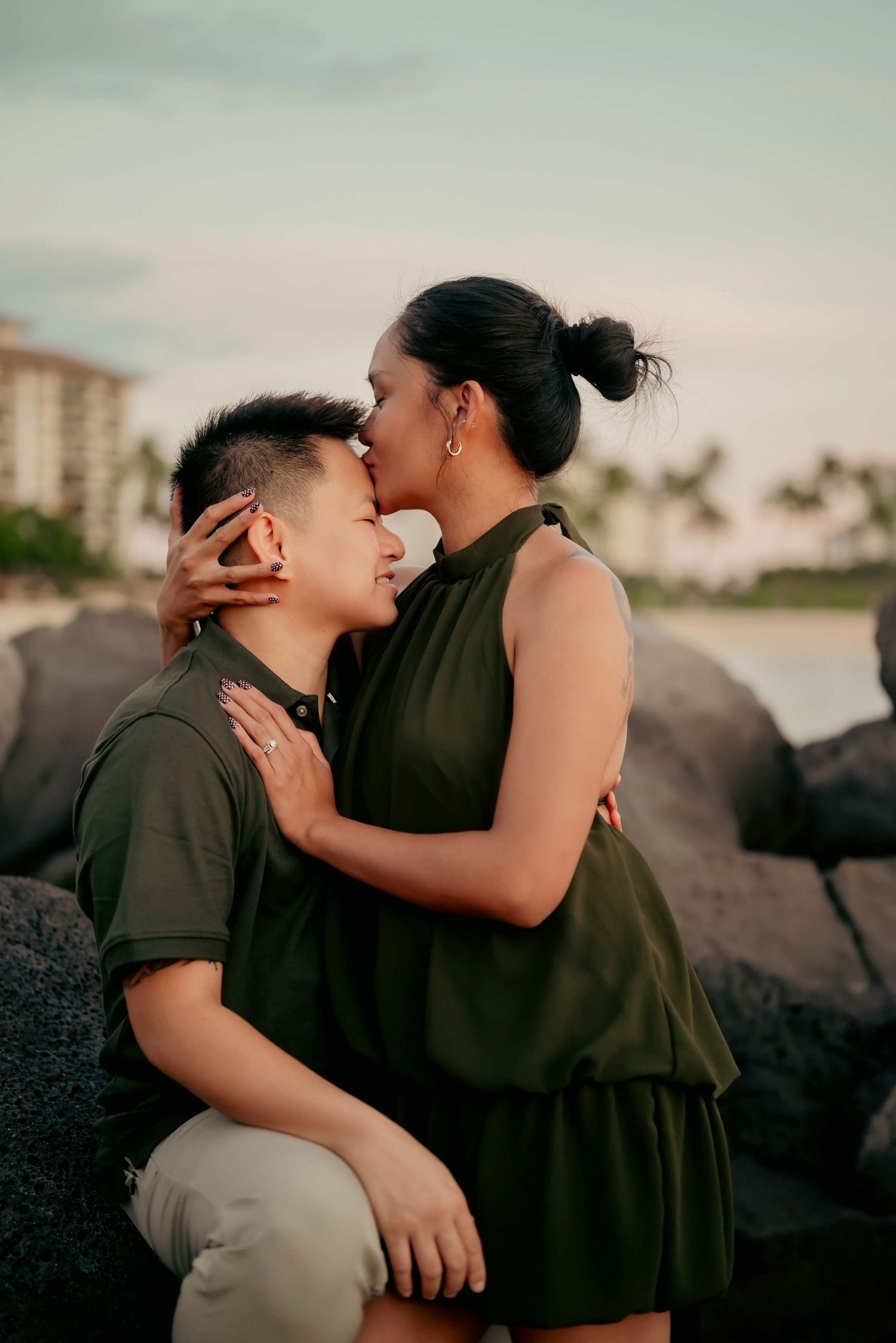 A romantic couple in olive green outfits share an intimate moment on rocky beach at sunset with palm trees in background.