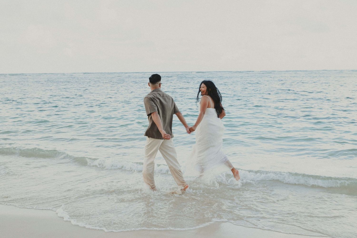 A couple in wedding attire runs joyfully along the shoreline with waves lapping at their feet during sunset.