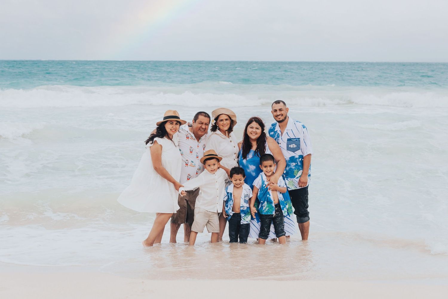 Family of seven poses on a sandy beach with ocean waves and a rainbow in the background.