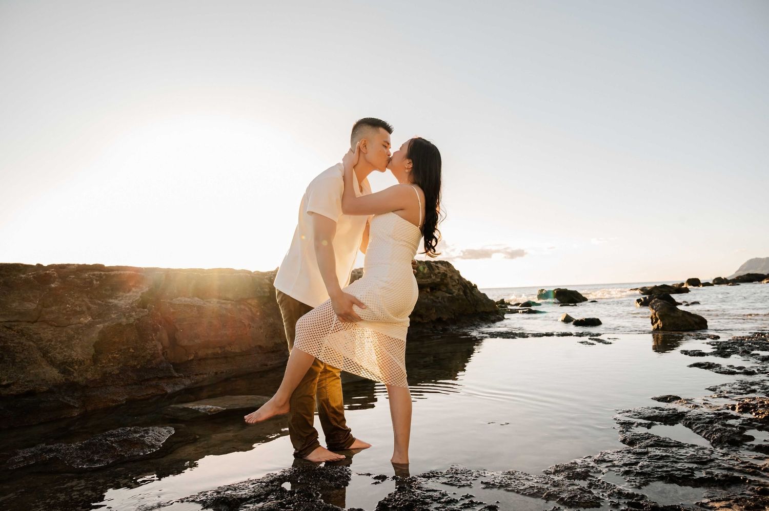 Couple sharing a romantic kiss at sunset on rocky beach, golden backlight illuminating their embrace by the ocean.