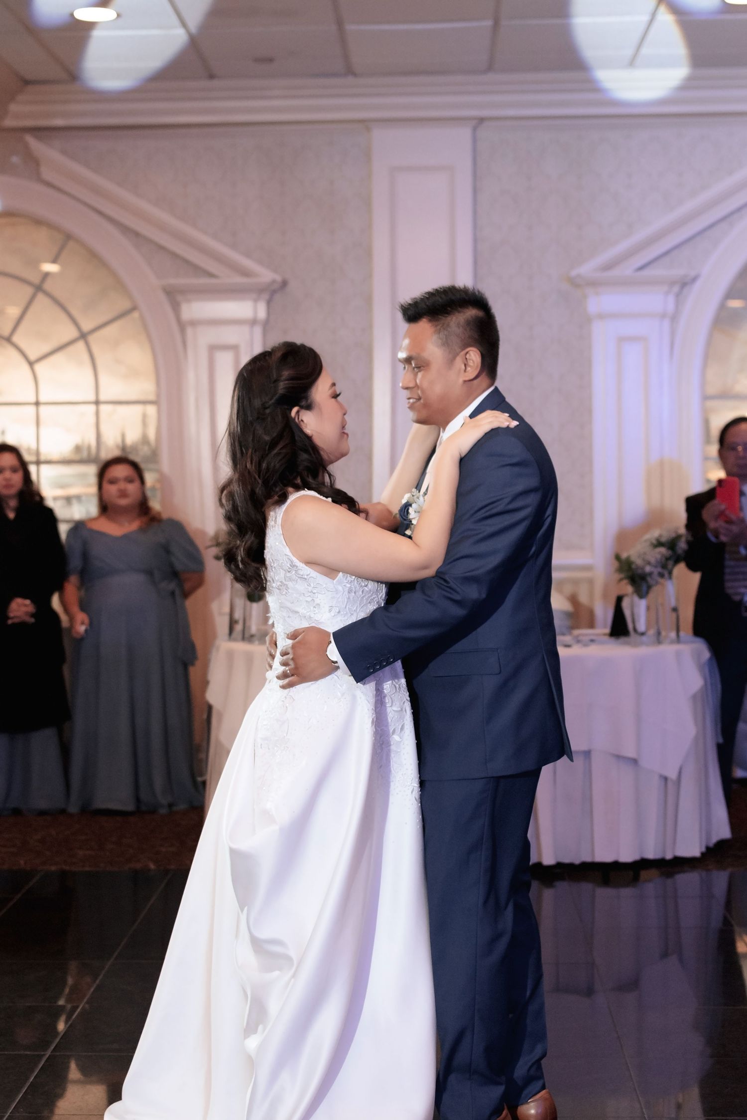 Bride and groom share their first dance at wedding reception in elegant ballroom with guests watching in background.