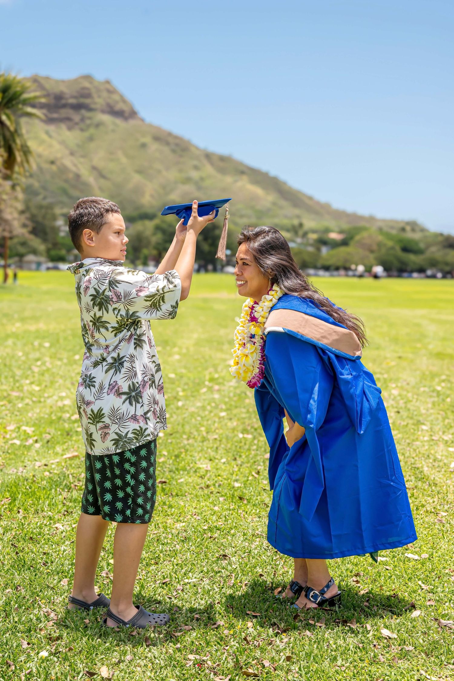 Boy placing graduation cap on woman in blue gown with lei, celebrating outdoors near Diamond Head, Hawaii.