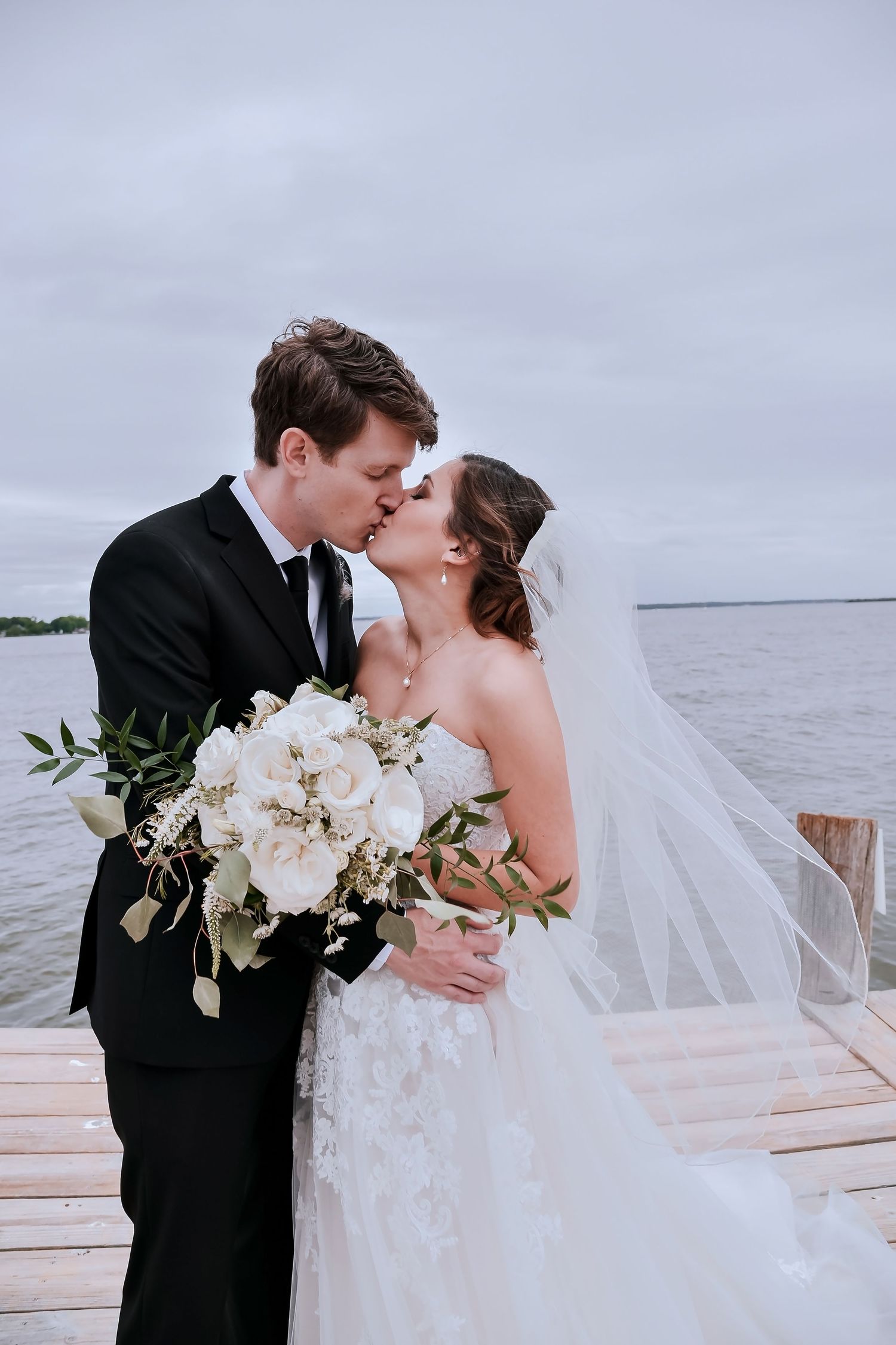 A newlywed couple shares romantic kisses on a dock overlooking a serene lake at sunset.