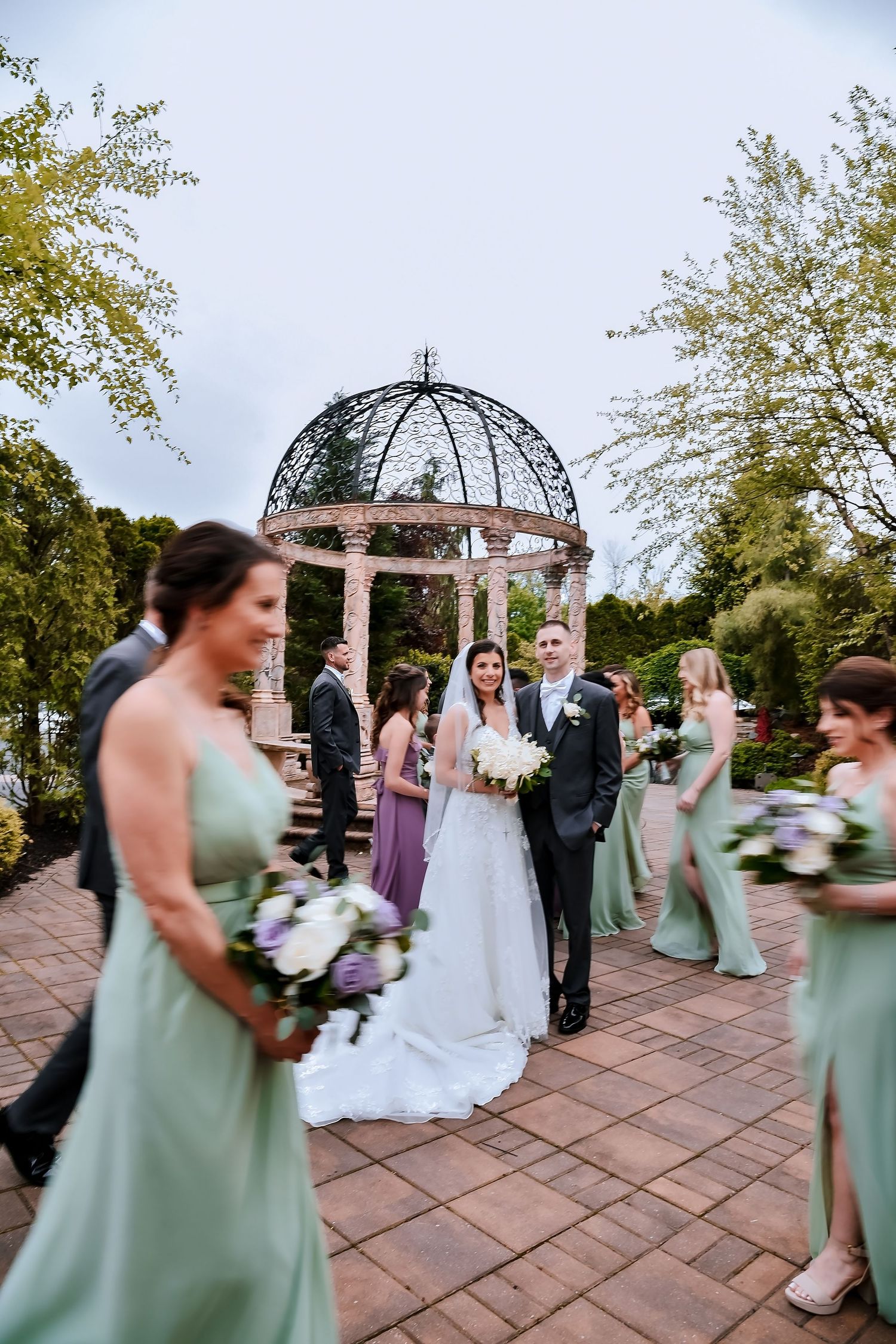Wedding ceremony taking place under an ornate metal gazebo with guests in sage green dresses on a brick patio.