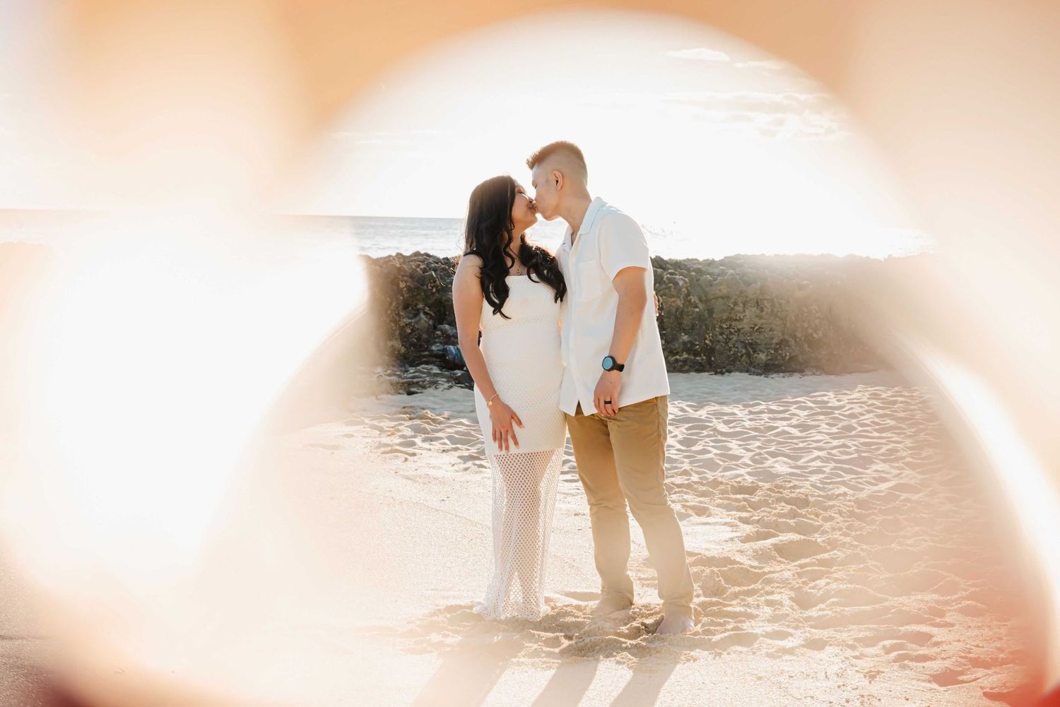 Couple sharing a romantic kiss on a sunlit beach, framed by a soft bokeh foreground with ocean waves behind them.