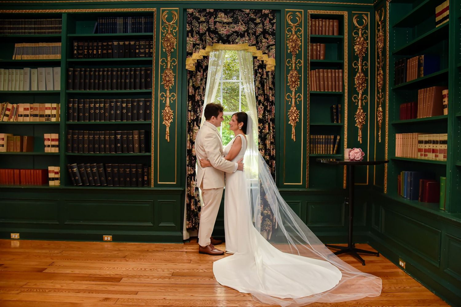 A couple embraces in an ornate emerald green library with floor-to-ceiling bookshelves and a flowing white wedding veil.