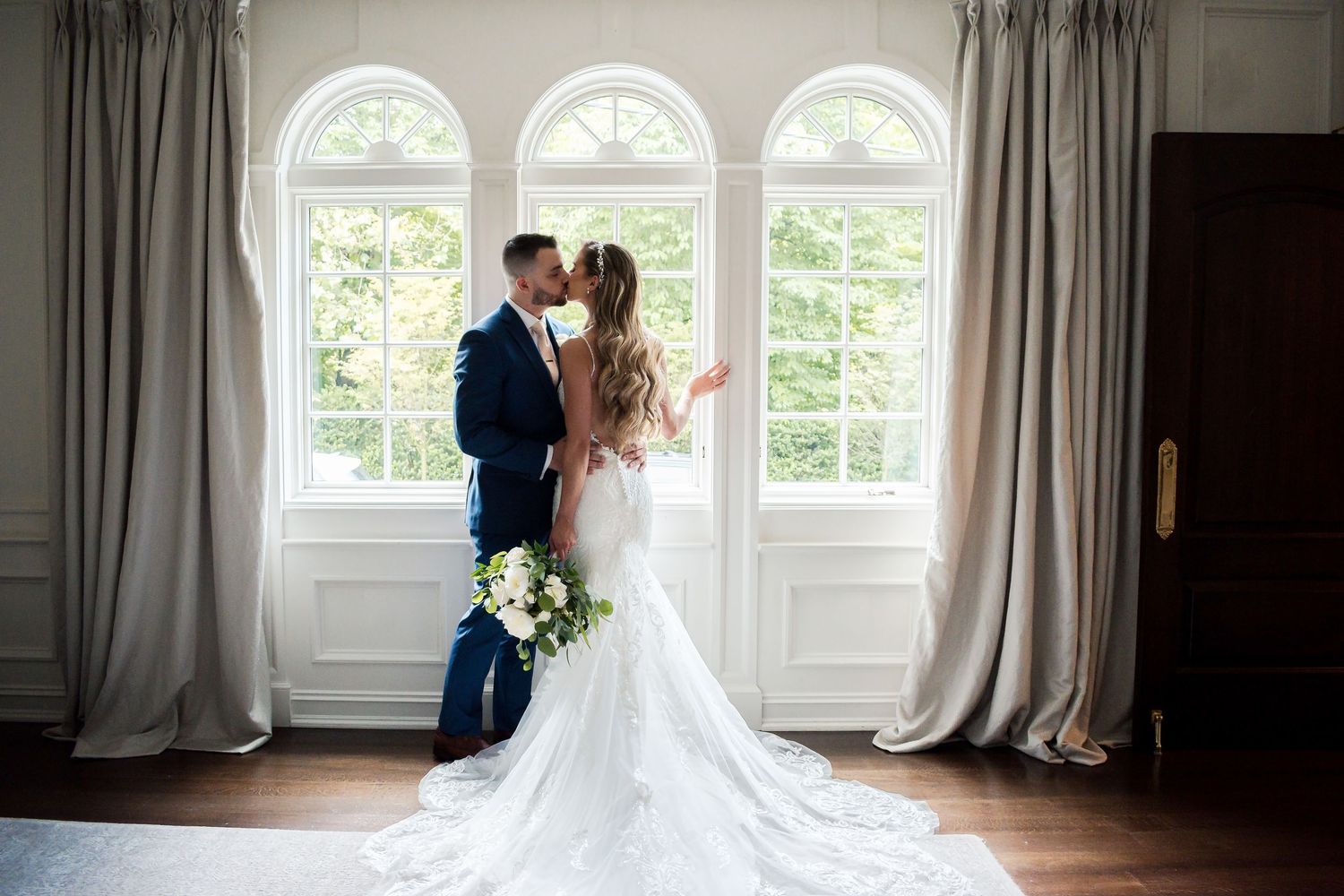 Elegant wedding couple sharing an intimate moment by grand arched windows with flowing curtains and a beautiful bridal bouquet.