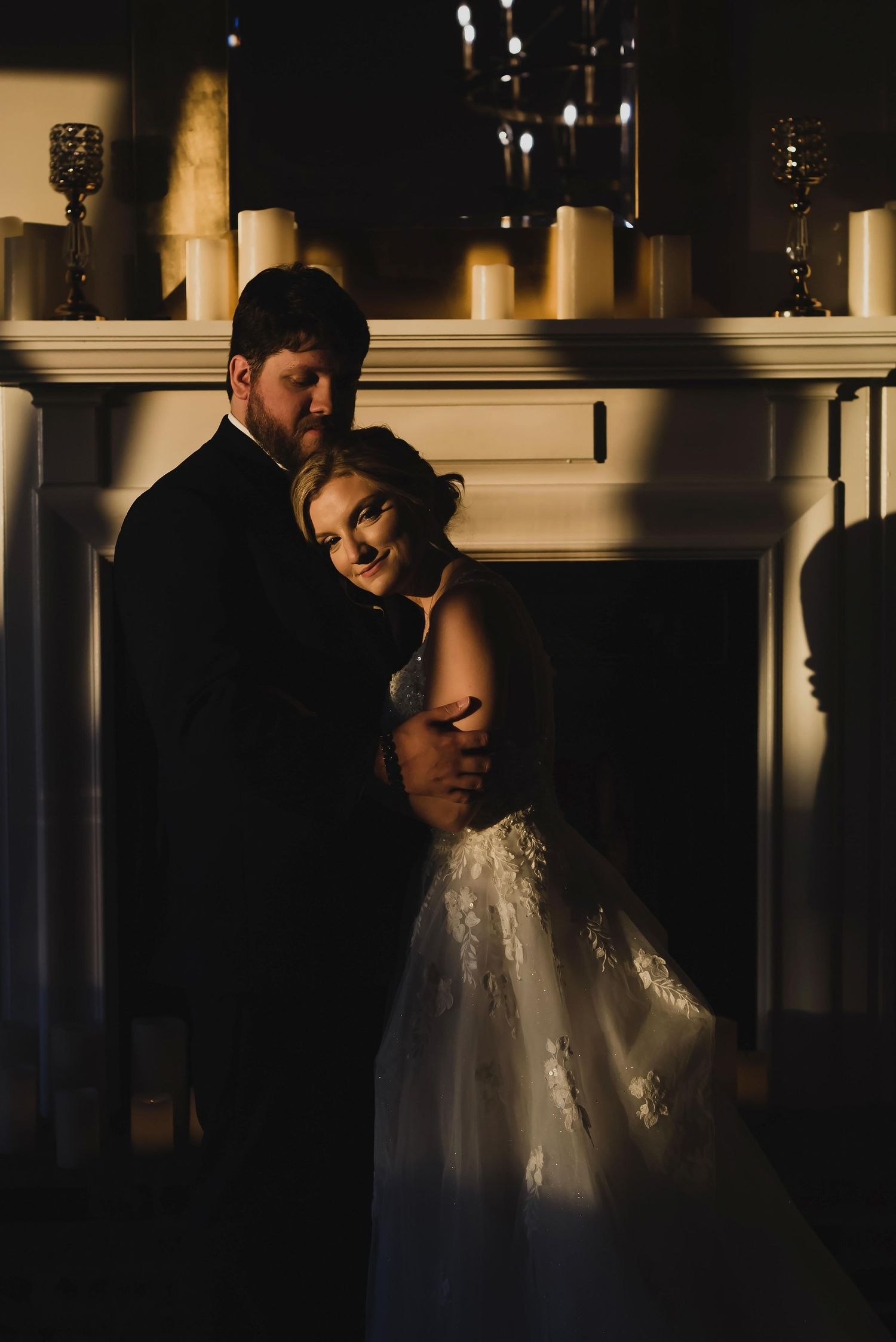 Dramatic wedding portrait in moody lighting shows couple embracing beside a fireplace with candles glowing above.