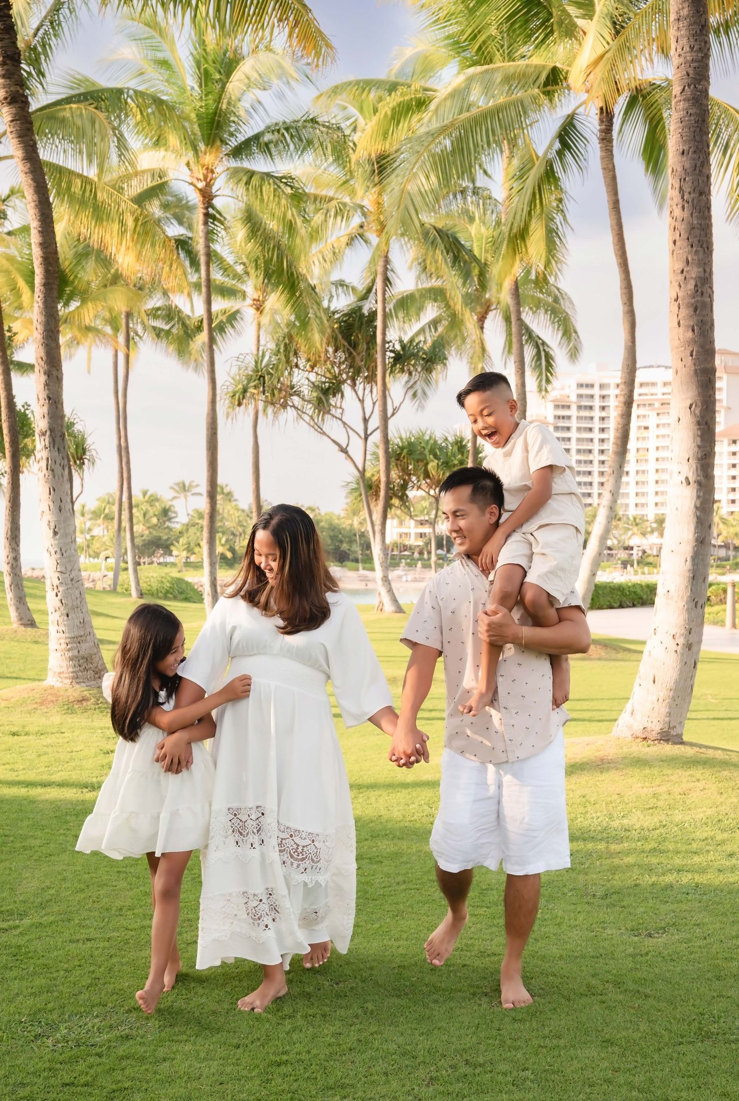 Family of four in white outfits laughing together on green lawn with tropical palm trees in Hawaii.