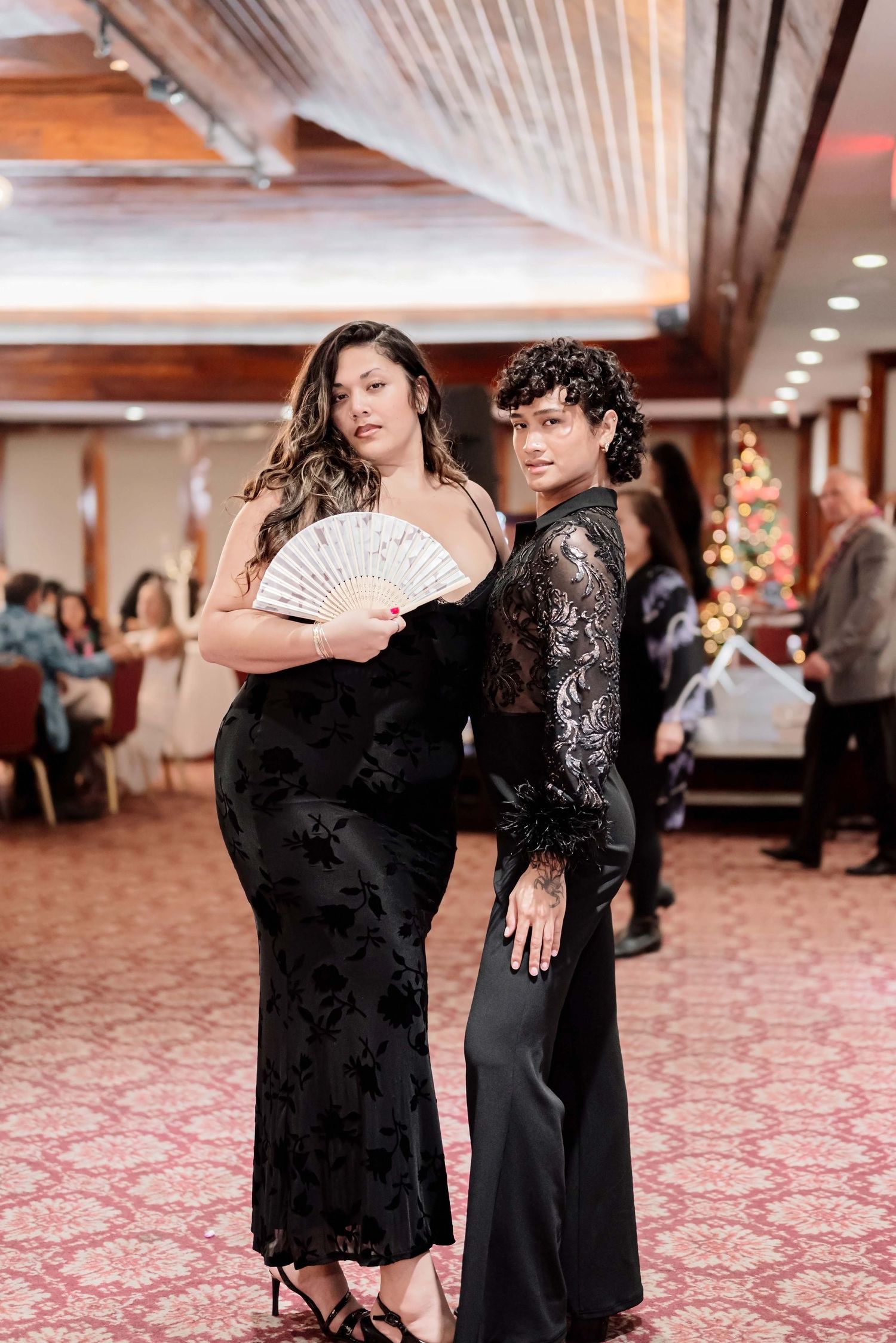 Two people in elegant black formal attire pose together at an indoor event, one holding a decorative white fan.