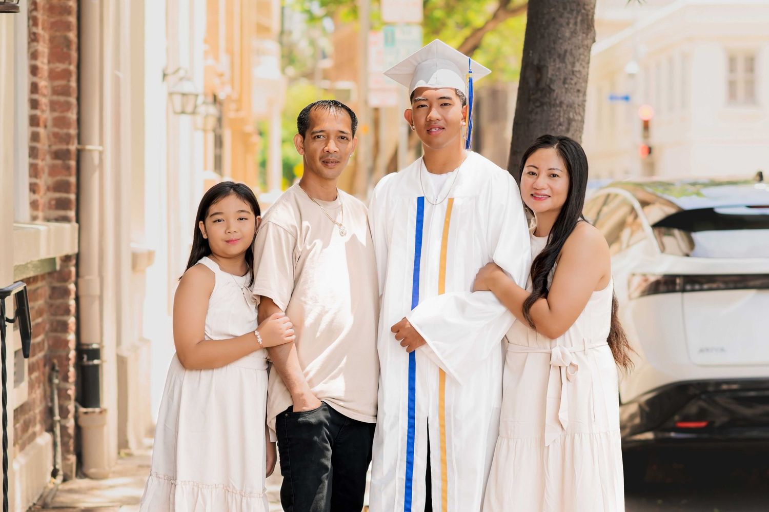Family of four in white outfits celebrating graduation outdoors, with graduate in cap and gown holding diploma.