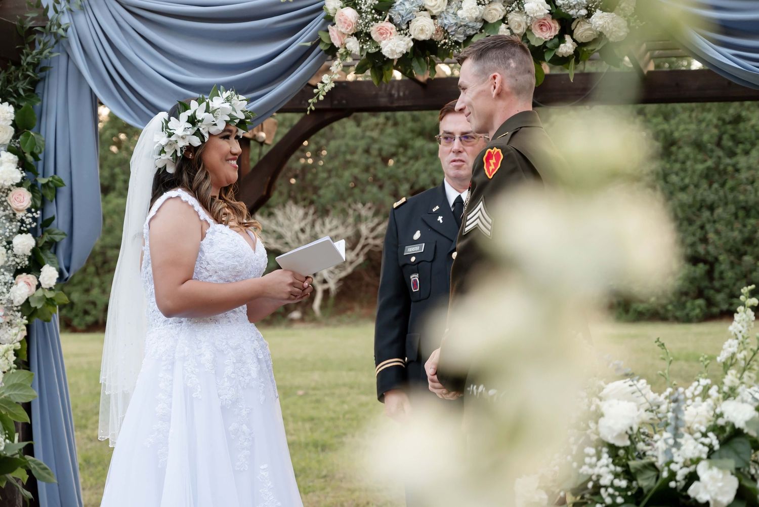 Military groom and bride in white gown exchange vows outdoors under floral arch with blue draping.