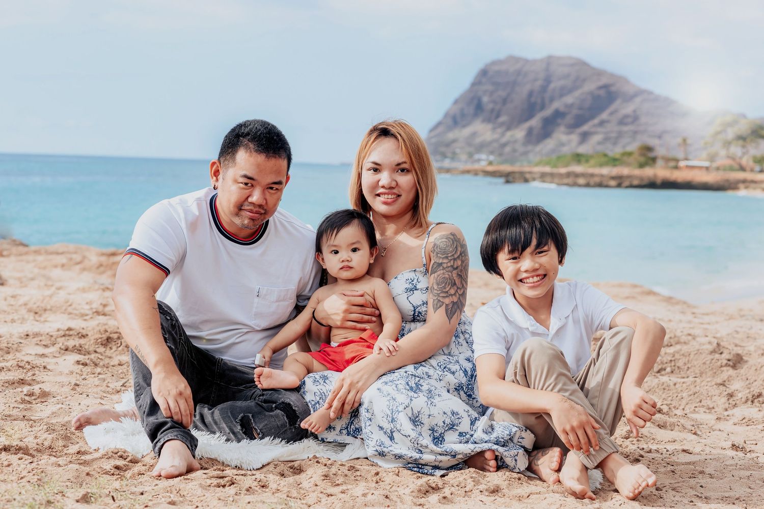 Family of four smiling together on a sandy beach with a scenic mountain and ocean backdrop on a sunny day.