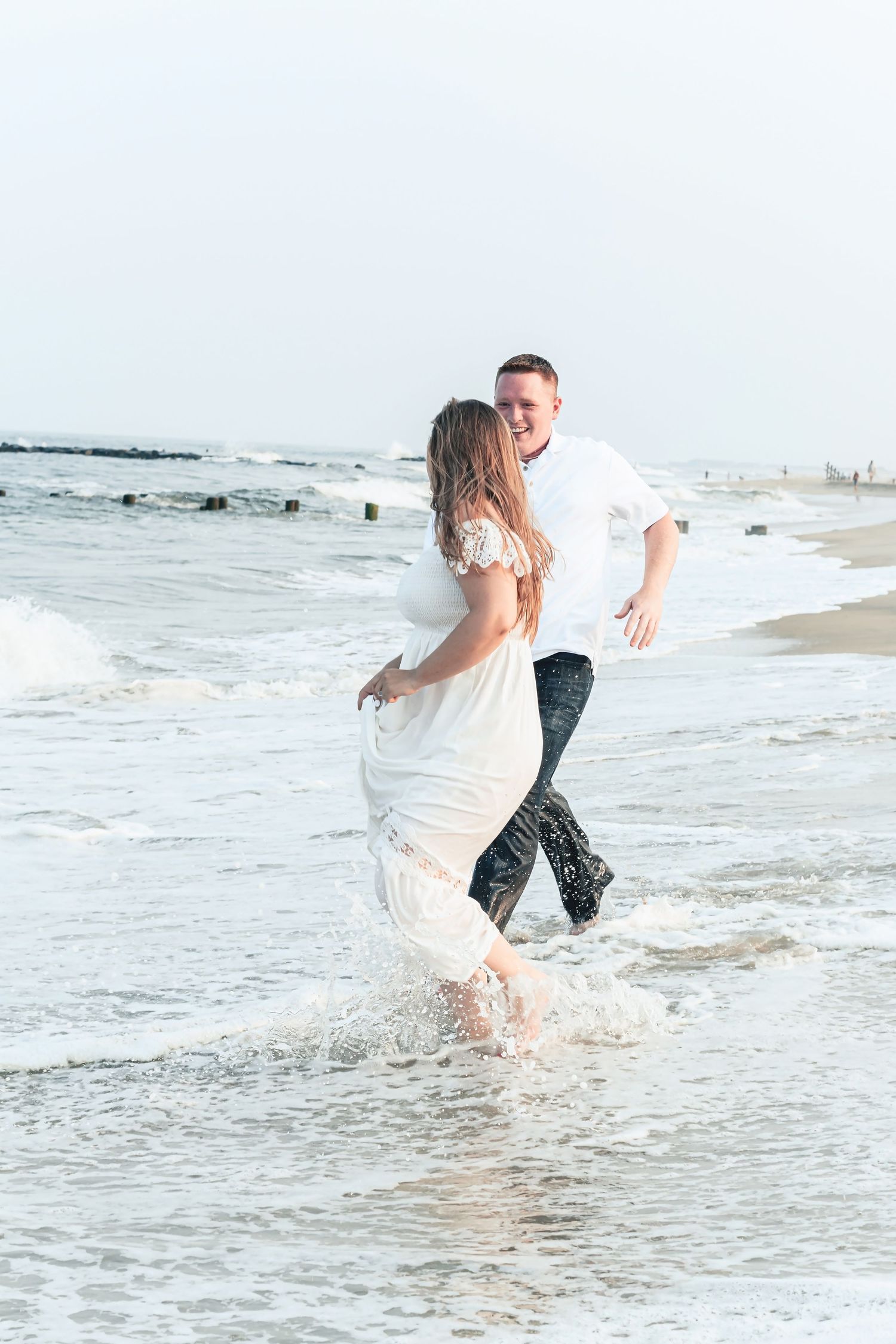 Couple in white wedding attire laughing and running through ocean waves on a sandy beach.