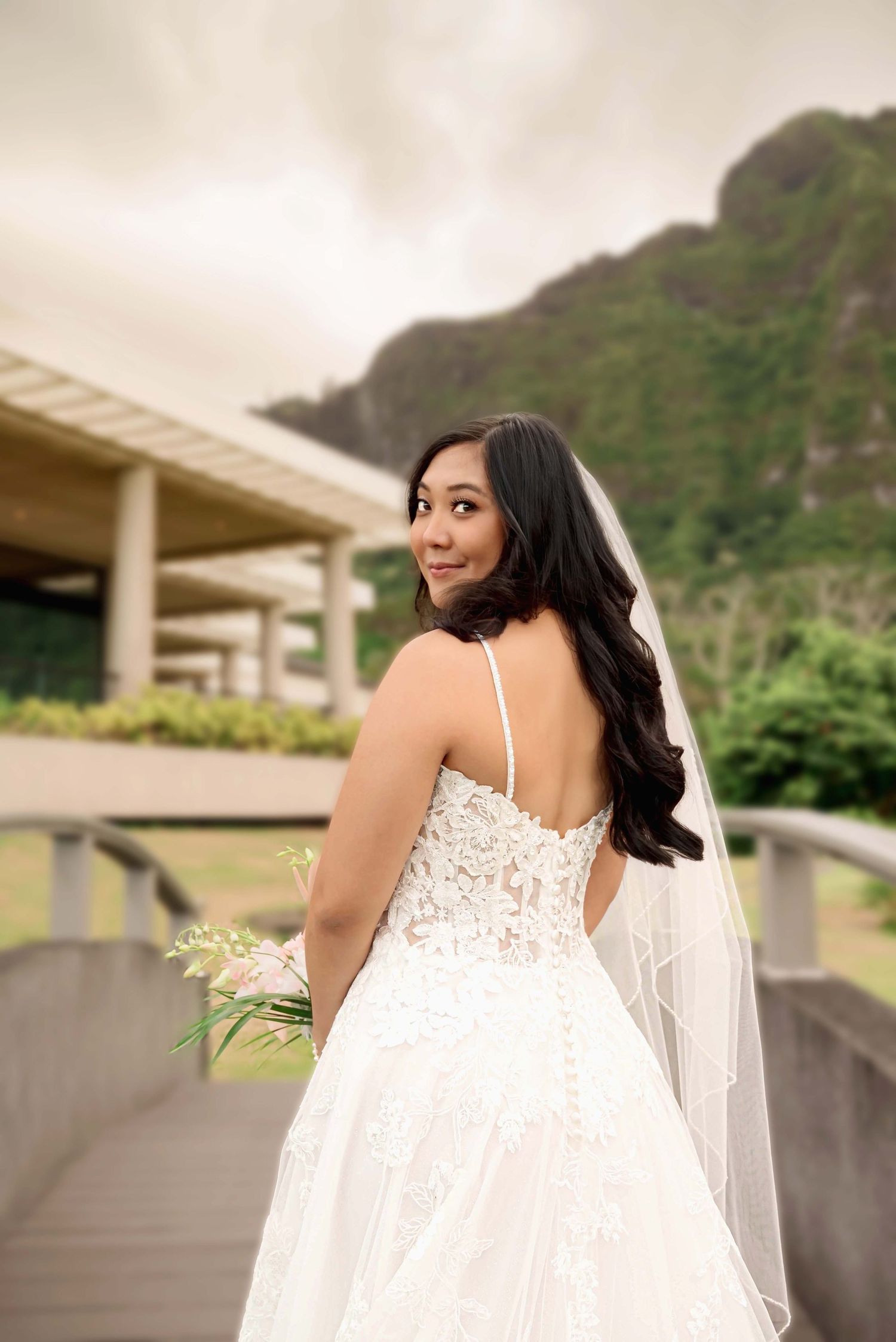 Bride in lace wedding gown and veil holds bouquet, posing on bridge with lush mountain backdrop.