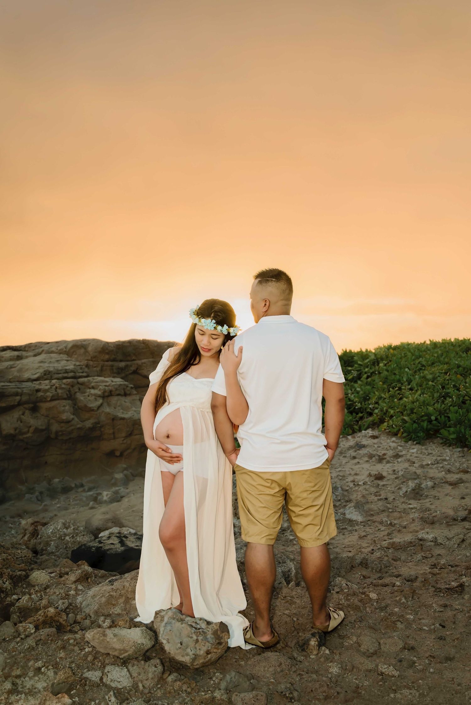 Pregnant woman in white sheer gown and flower crown stands with partner at sunset on rocky Hawaii coastline.
