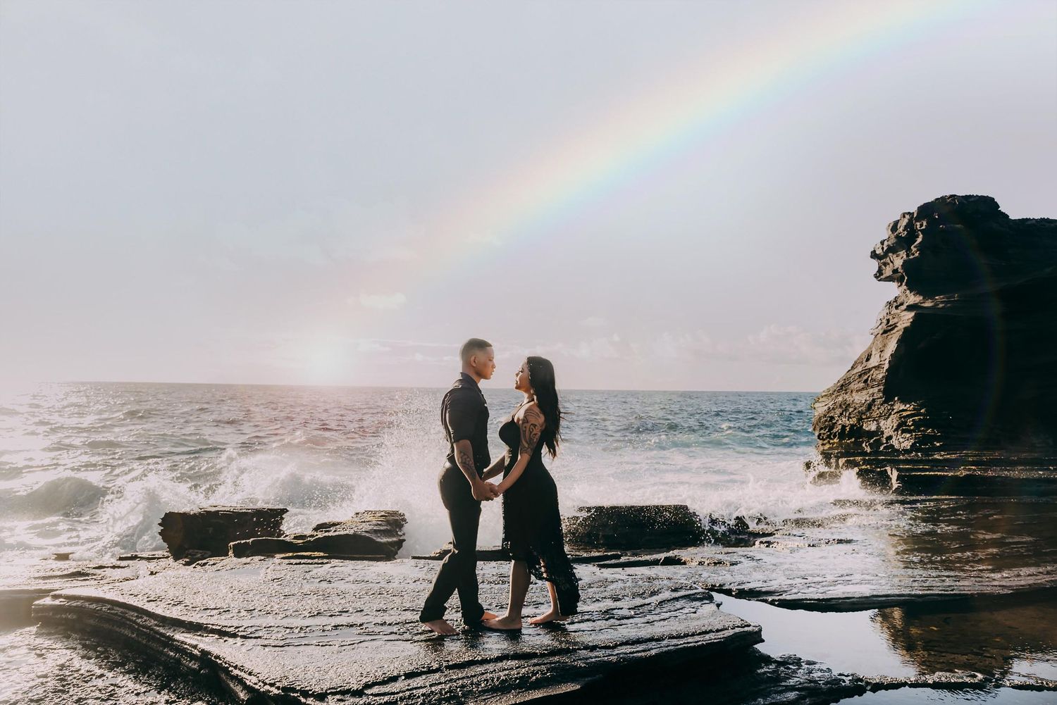 Two silhouettes stand together on a rocky beach at sunset with a rainbow arching across the misty sky.