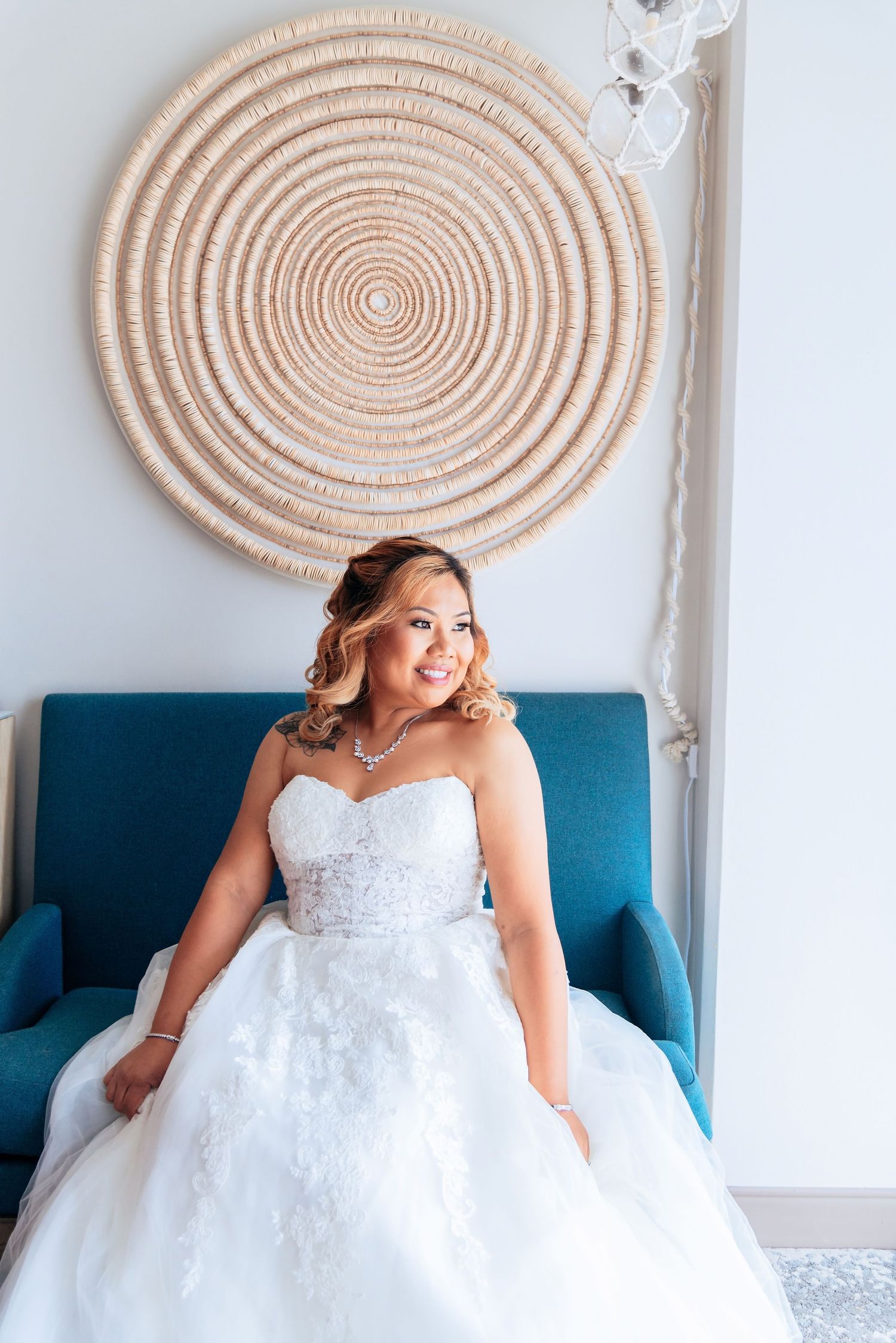Smiling bride in a strapless white ball gown sits in a teal chair before a circular woven wall art.