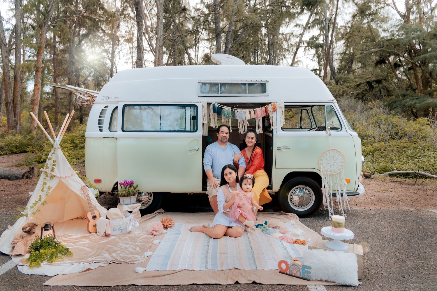 Family posing with a vintage mint green VW bus, teepee, and picnic setup outdoors in a forest setting.