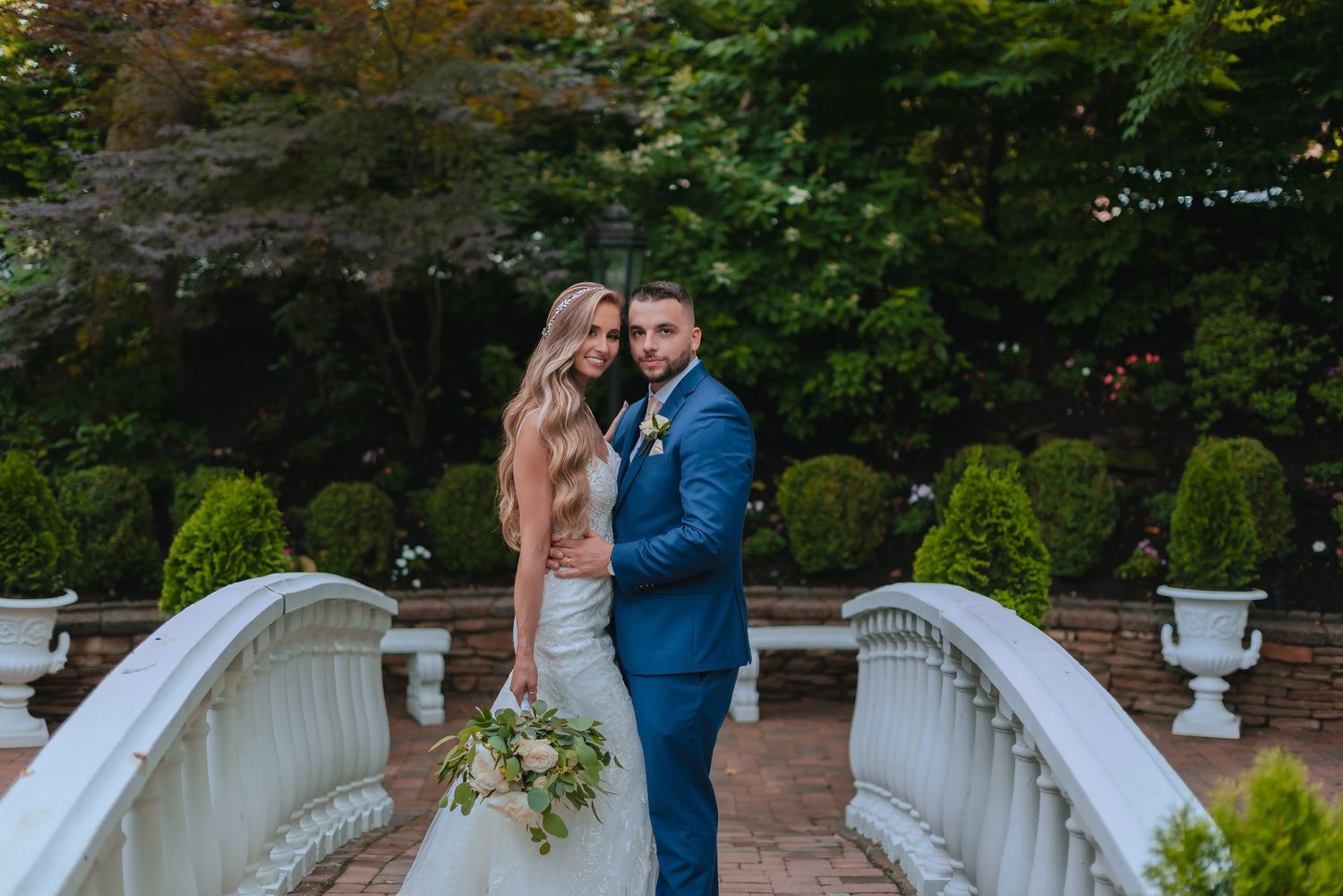 Bride and groom pose on a white bridge surrounded by lush greenery and topiaries at an elegant garden wedding venue.