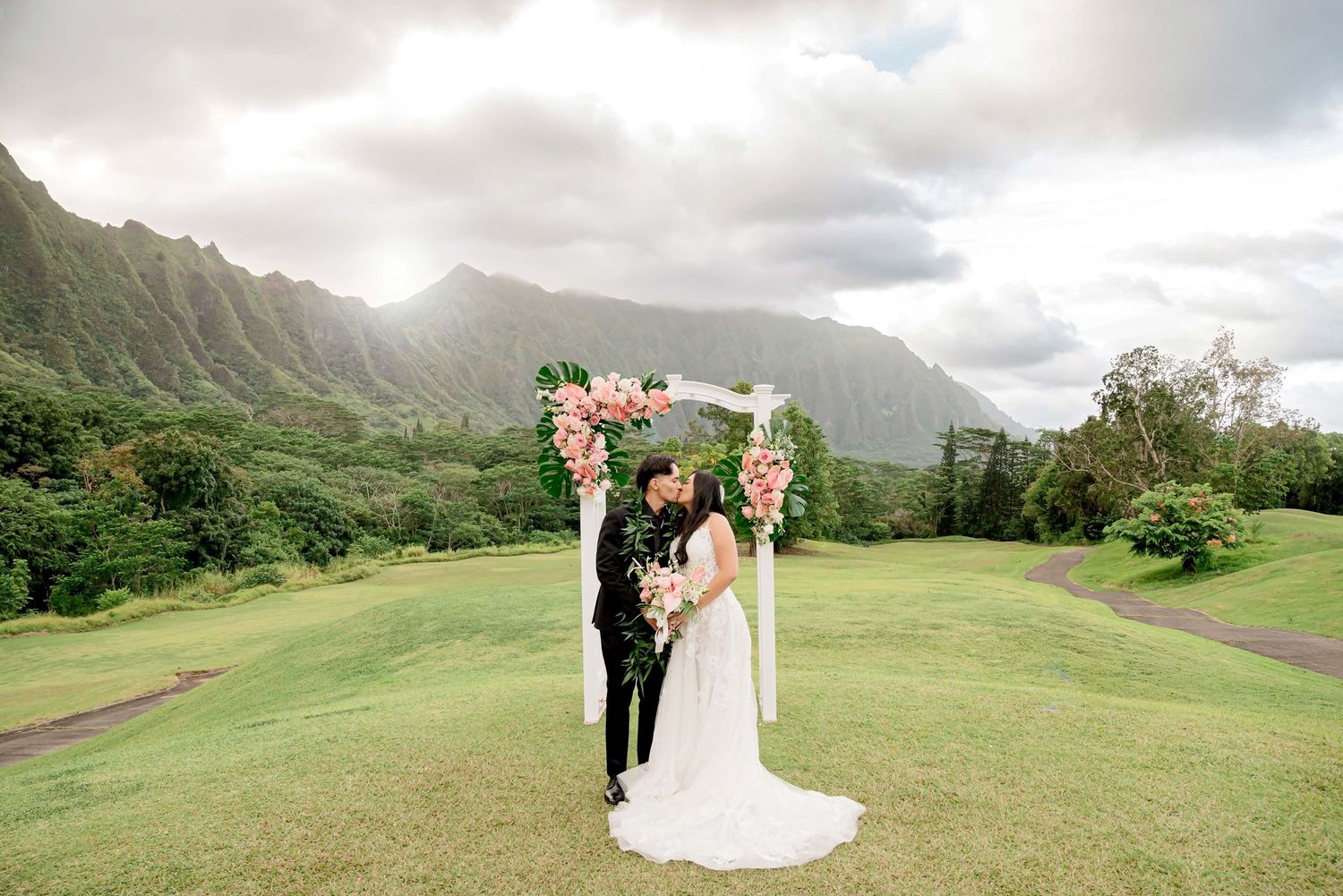 A couple poses for wedding photos on a scenic golf course with dramatic mountain peaks and cloudy skies in Hawaii.