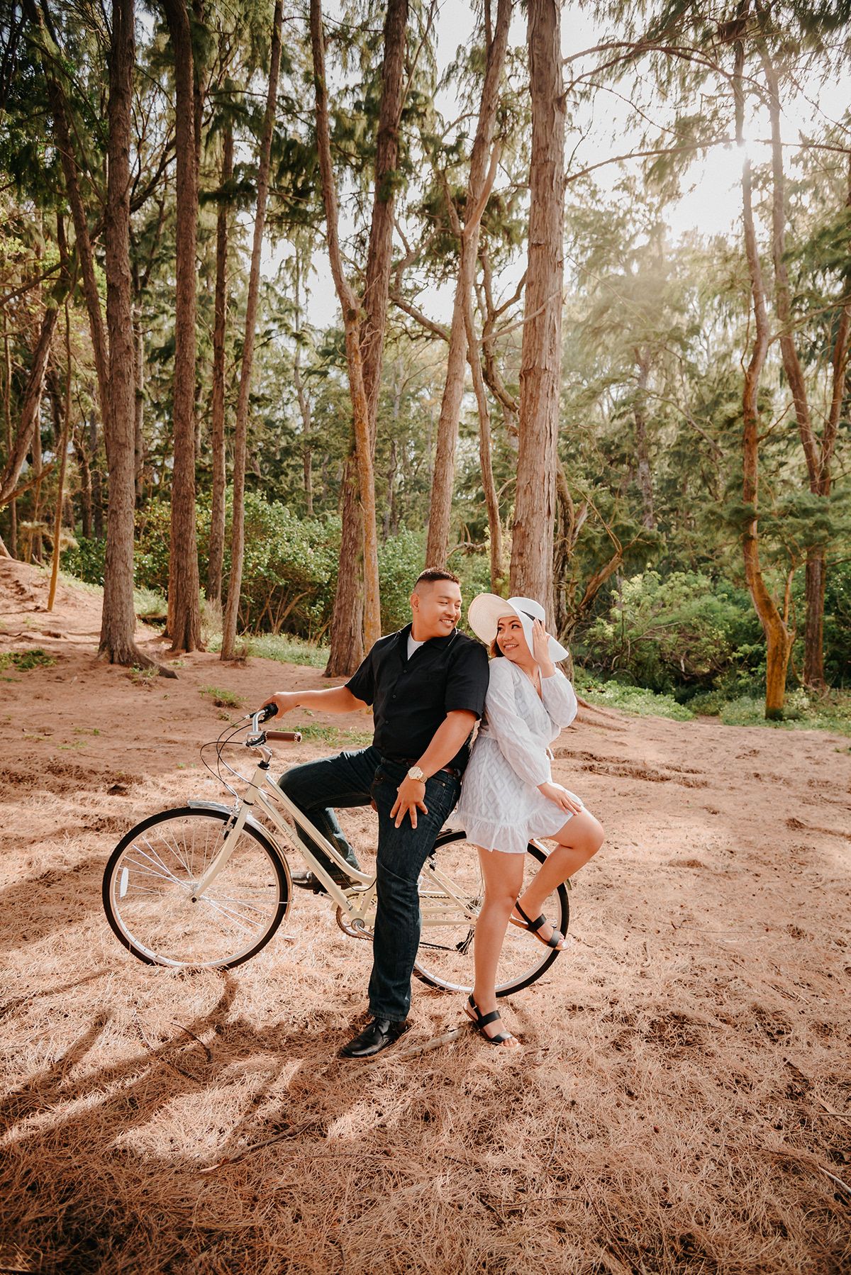 A couple poses playfully with a vintage bicycle in a sunlit forest setting with tall pine trees.