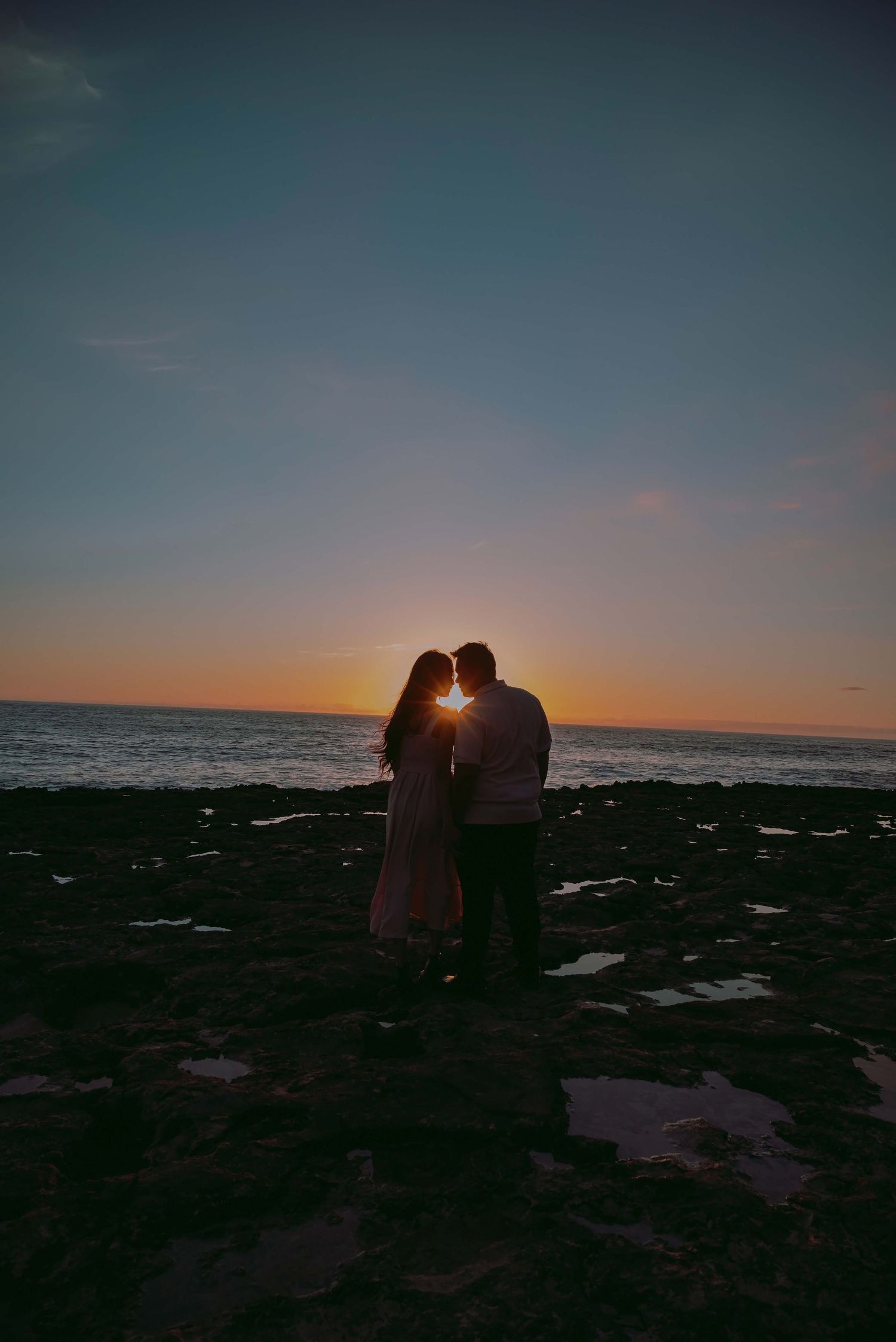 Silhouettes of a couple embracing against a dramatic sunset sky at the beach with sun peeking through between them.