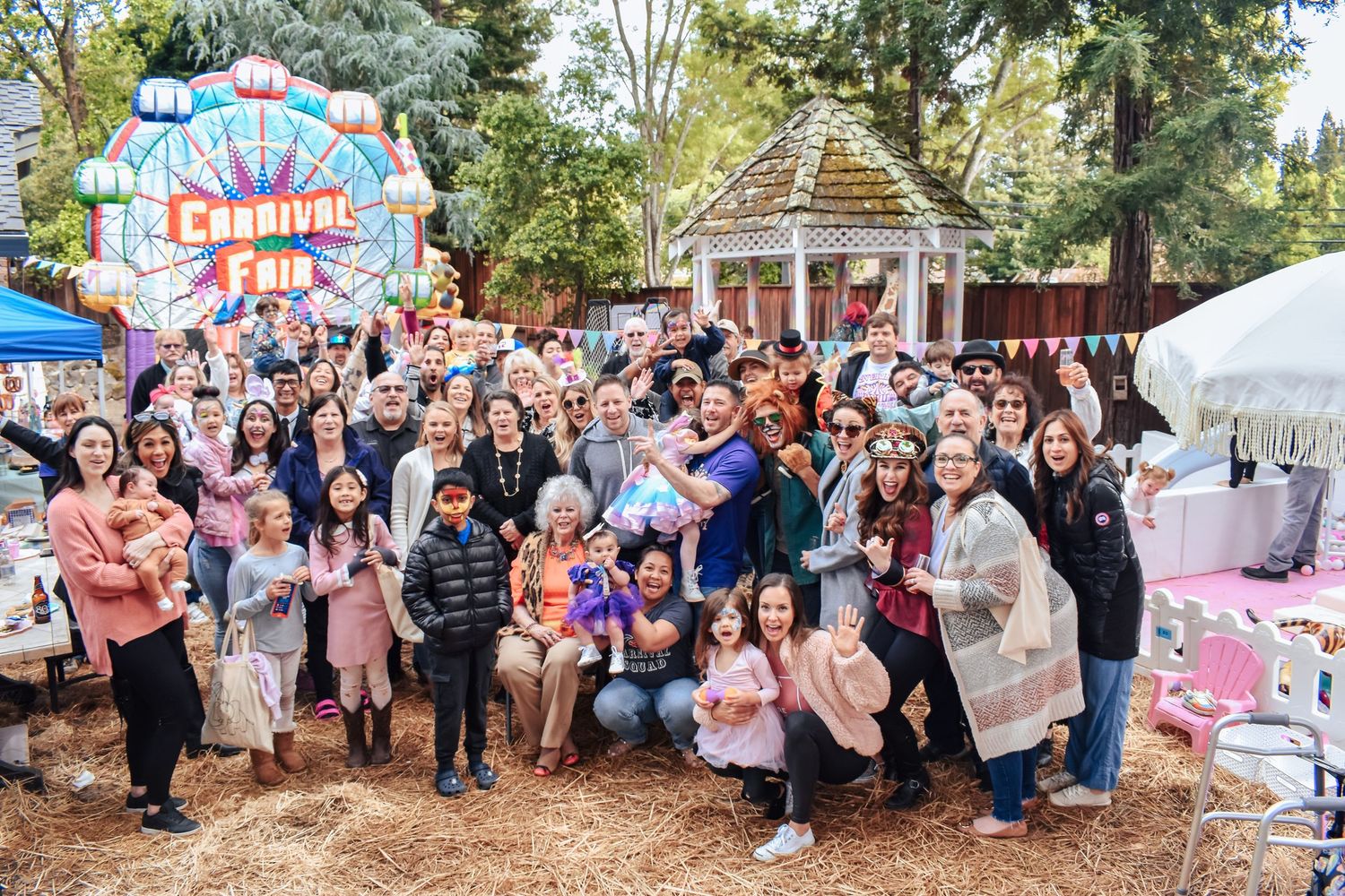A large crowd gathers outdoors for a festive carnival event with colorful decorations and a gazebo in the background.