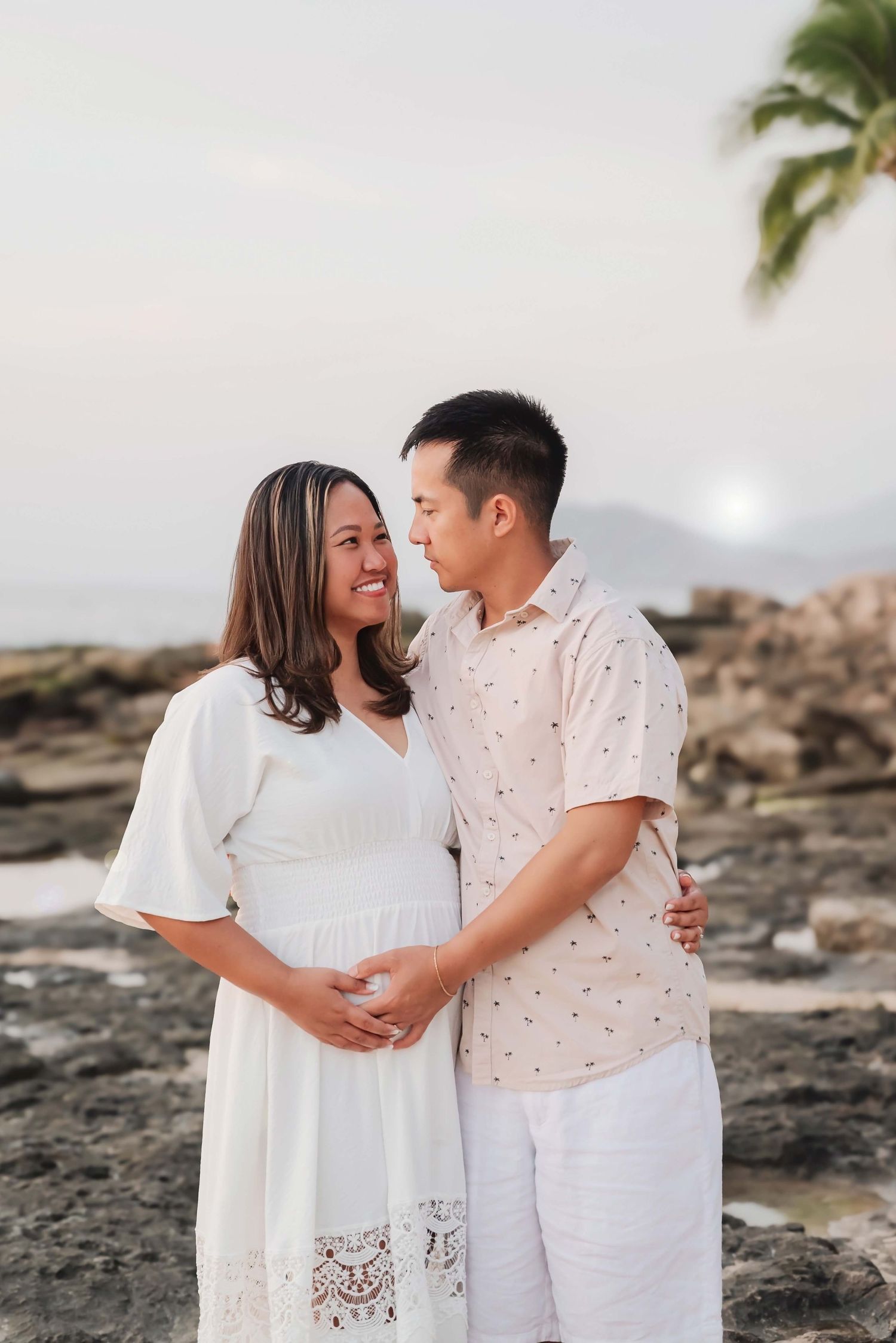 Pregnant couple embracing on rocky beach at sunset, woman in white dress smiling, man in floral shirt, tropical setting.