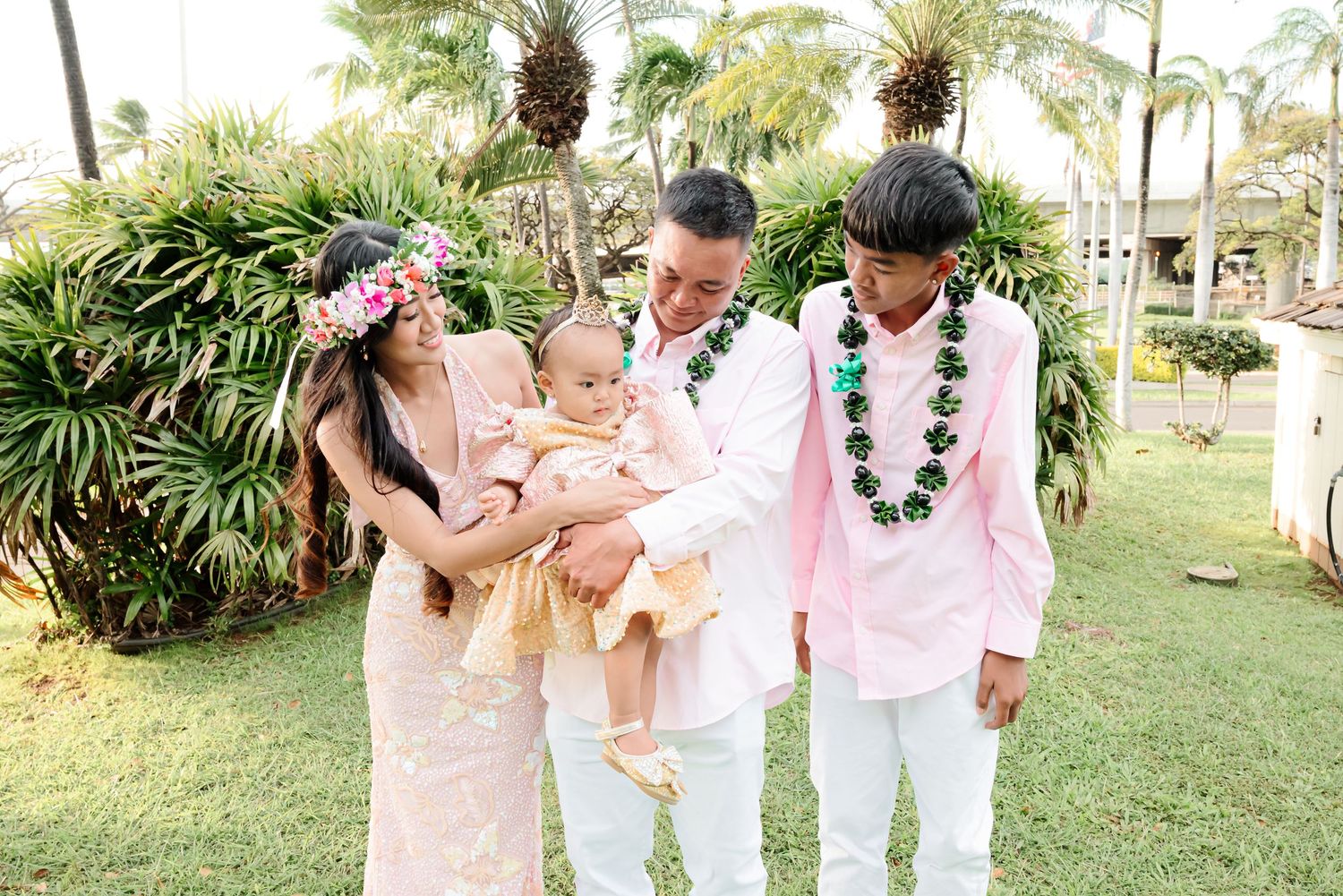 Family in Hawaiian attire with flower leis celebrating outdoors on green lawn with tropical palm trees.