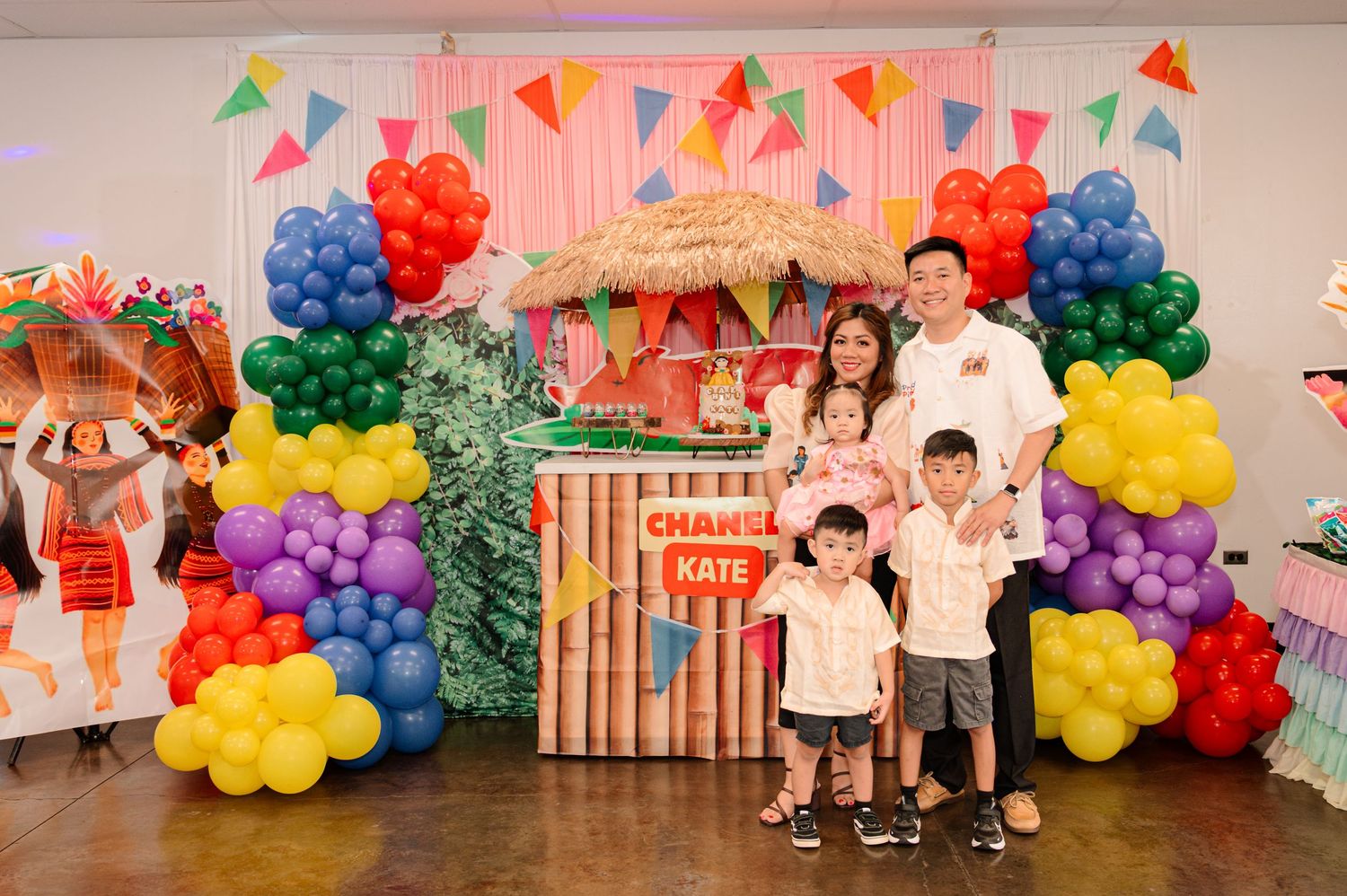 Family posing in front of colorful balloon arch at a Chanel and Kate birthday party with tropical tiki bar decor.