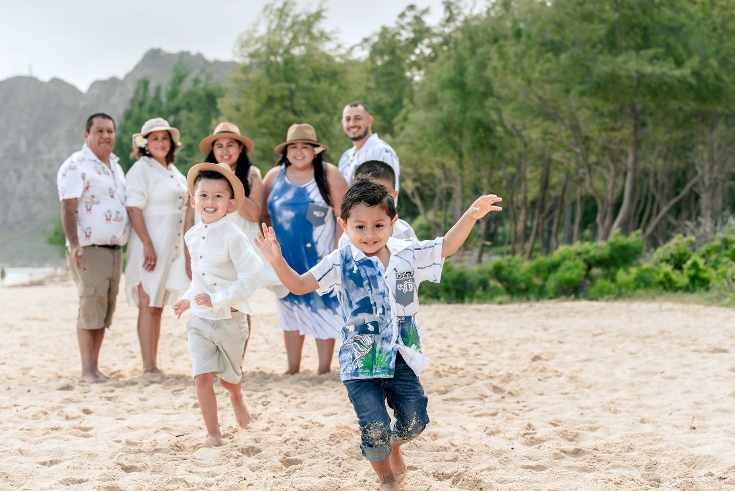 Family group photo on a sandy beach in Hawaii with two young boys running in the foreground and mountains behind.