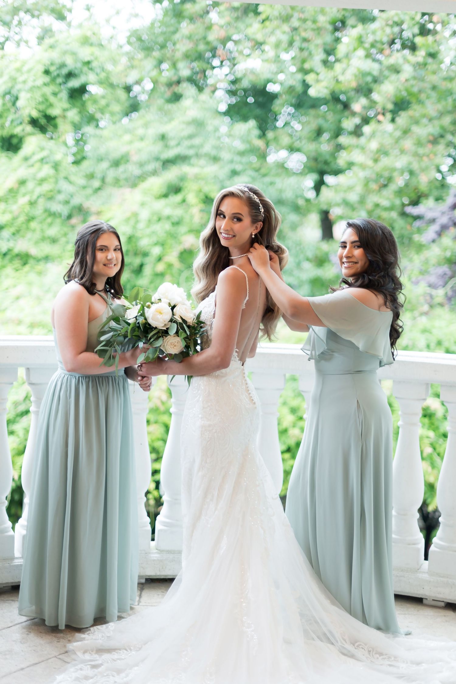 Bride and bridesmaids in sage green dresses pose together on a balcony with white bouquet and lush greenery backdrop.