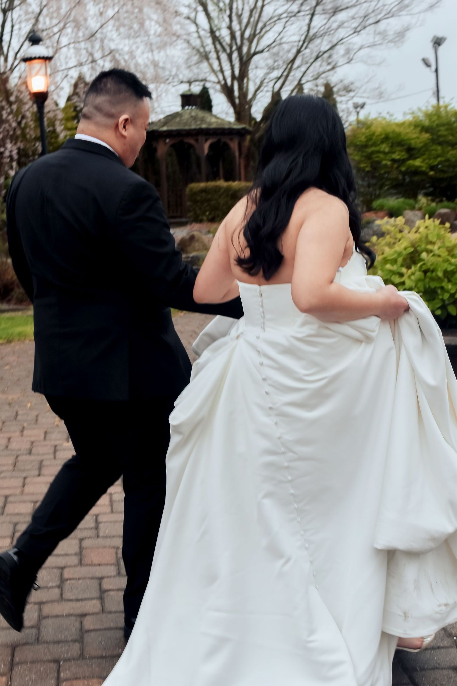 A bride in a white wedding gown walks alongside someone in a black suit on a brick pathway in a garden setting.